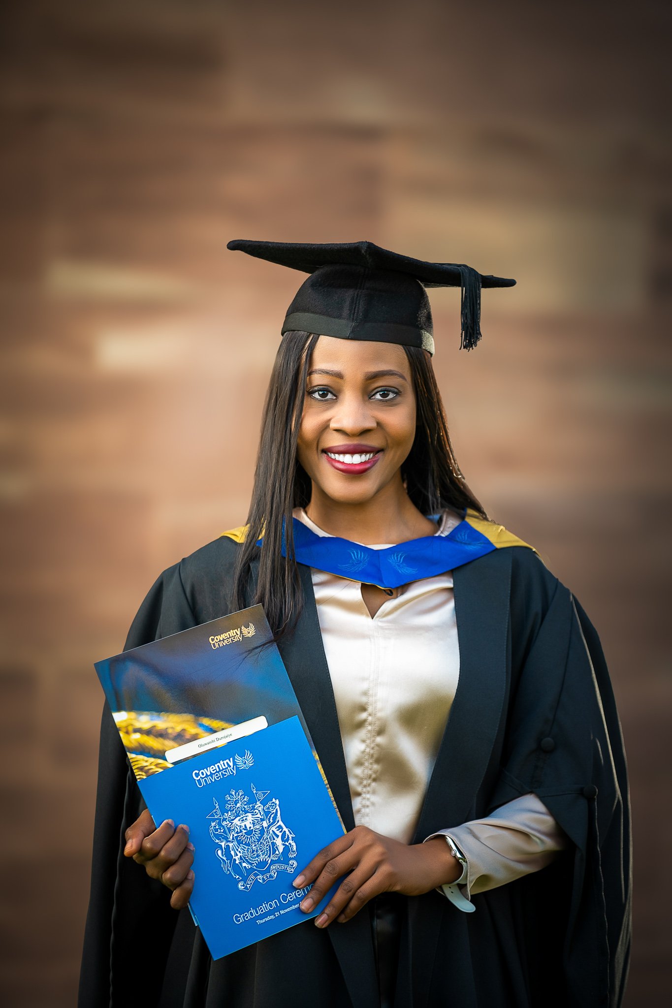 A woman in graduation gown and cap holding a diploma folder at a graduation ceremony.