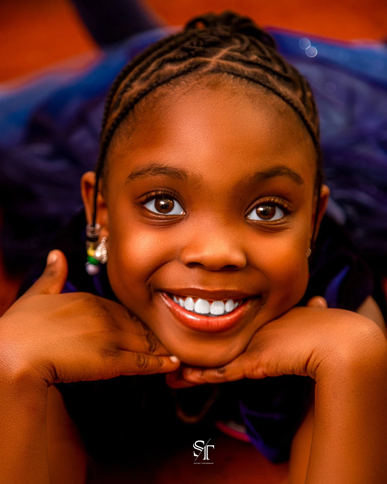 Close-up of a smiling young girl with braided hair and earrings, resting her chin on her hands, looking directly at the camera.