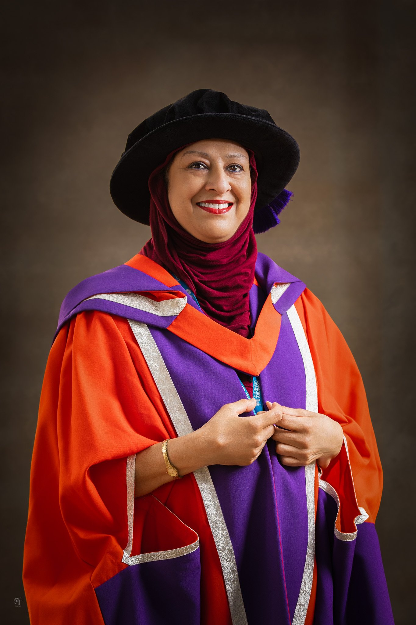 A woman wearing a graduation gown and cap, with a hijab, is smiling and holding a graduation medal, posed against a dark background.