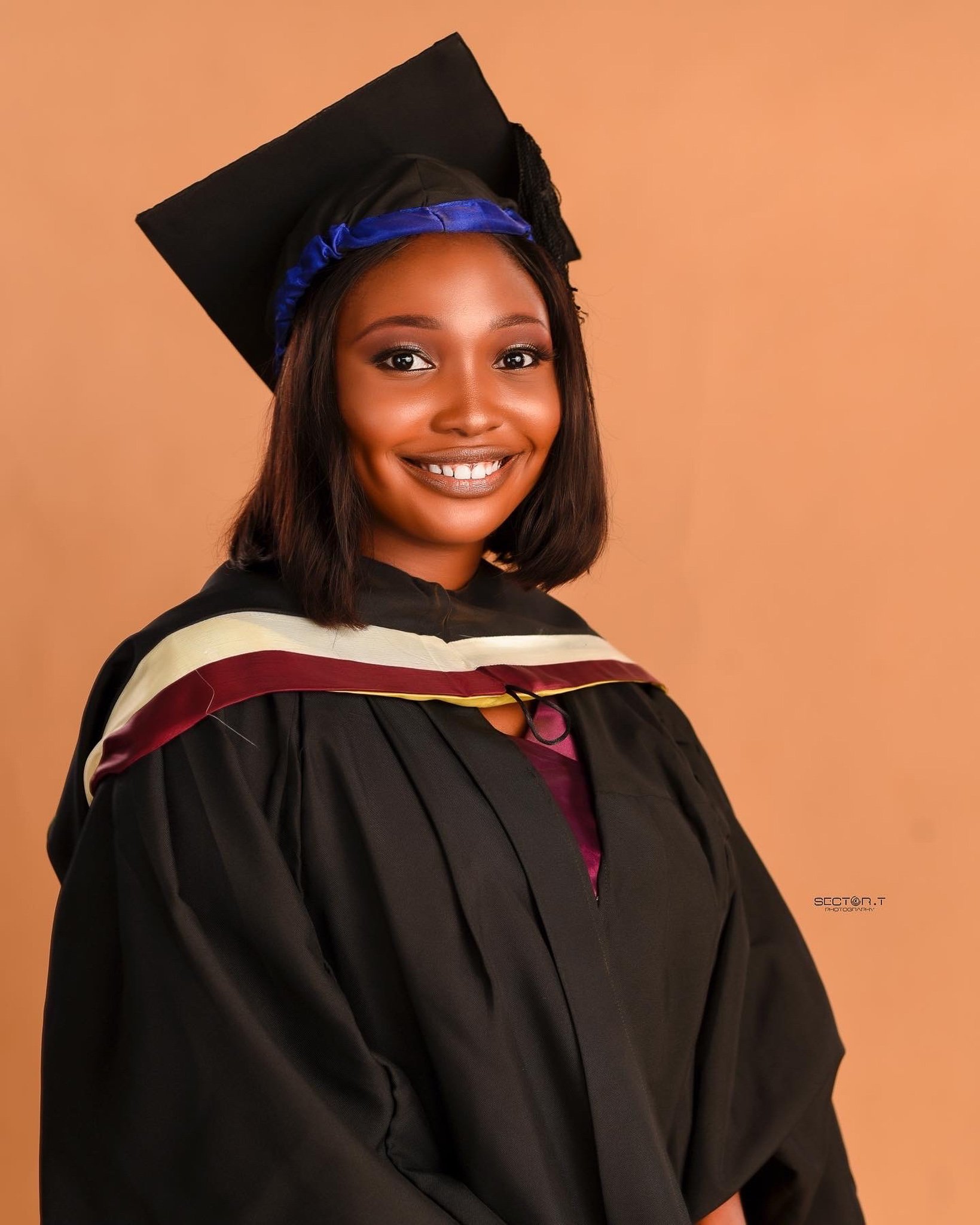 A young woman in a graduation gown and cap smiling at the camera, standing against a plain peach background.