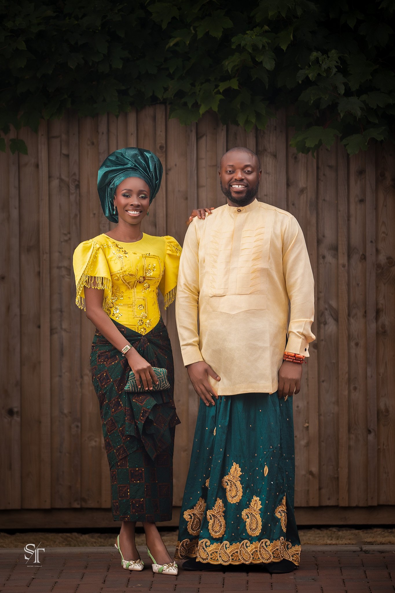 A man and woman standing together smiling in front of a wooden fence, the woman wearing a yellow traditional dress with headwrap and the man wearing a beige traditional shirt and blue patterned pants.