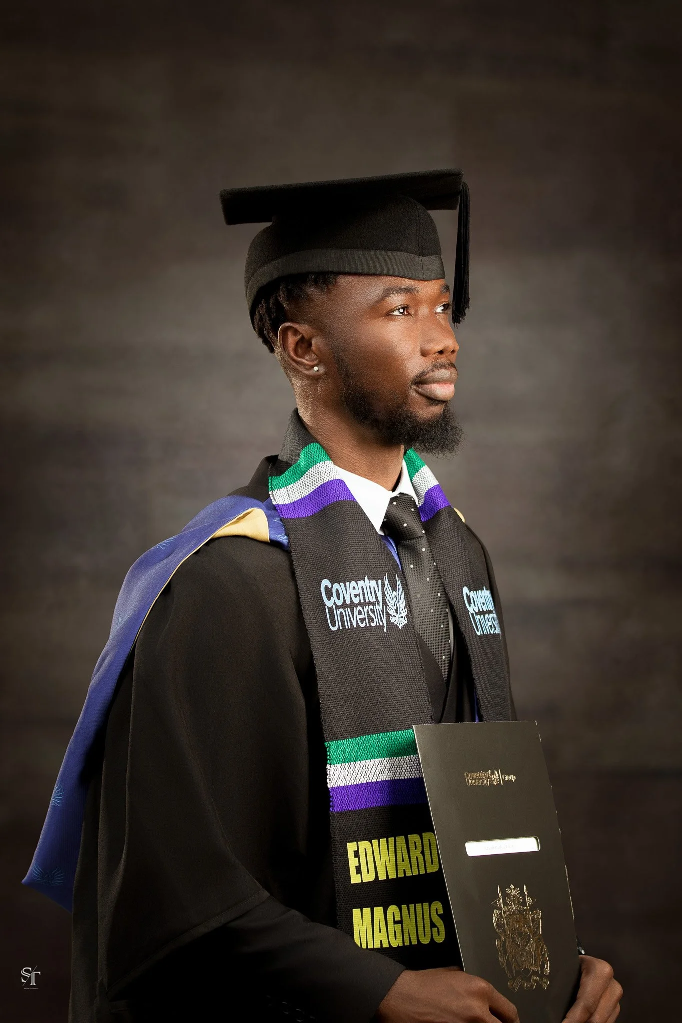 A young man in a graduation cap and gown holding a diploma and a program booklet, wearing a colorful stole, with a dark neutral background.