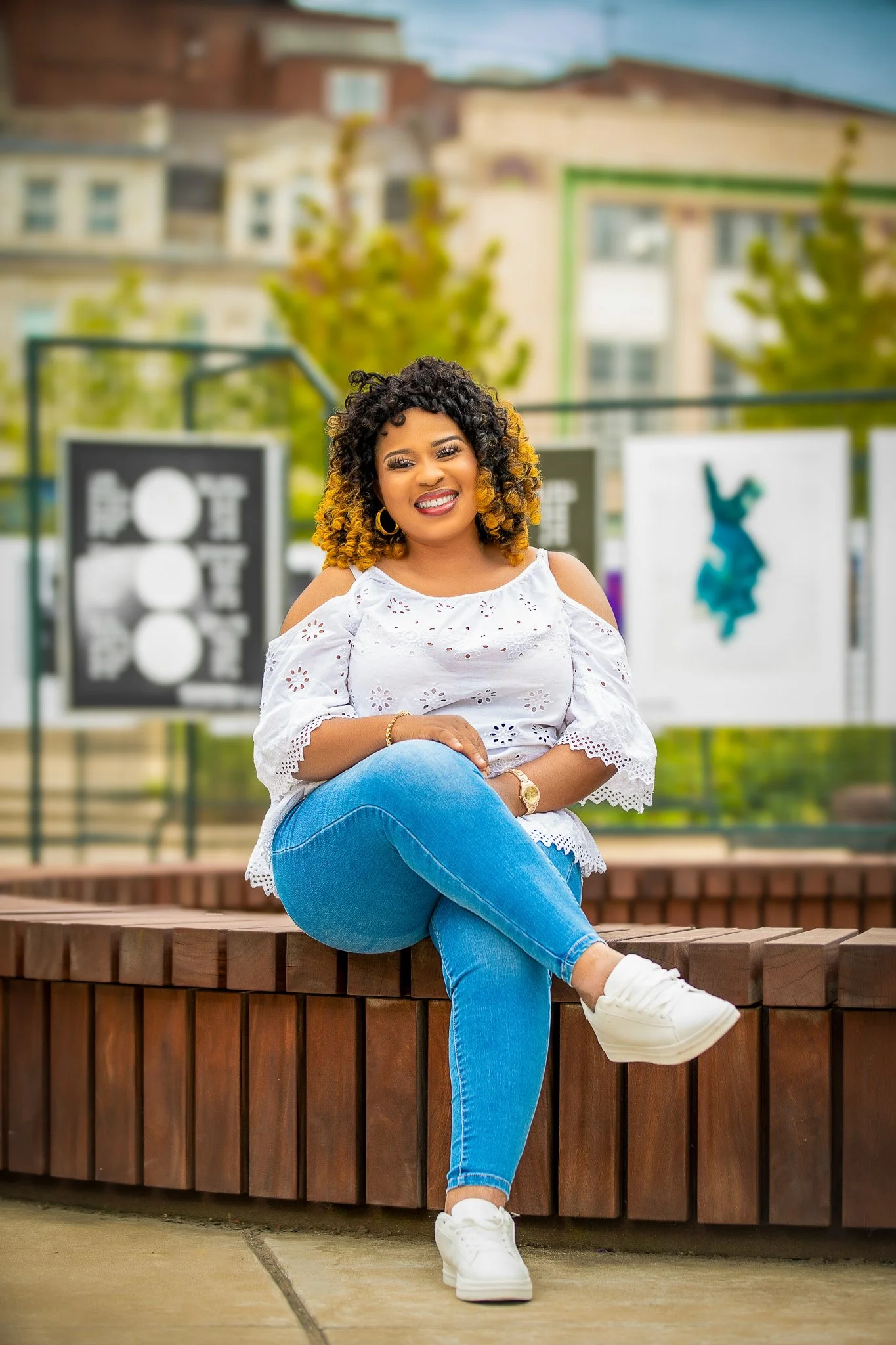 A smiling woman with curly, black and yellow hair, wearing a white embroidered off-shoulder top, blue jeans, and white sneakers, sitting on a wooden bench outdoors with colorful background art and buildings behind her.