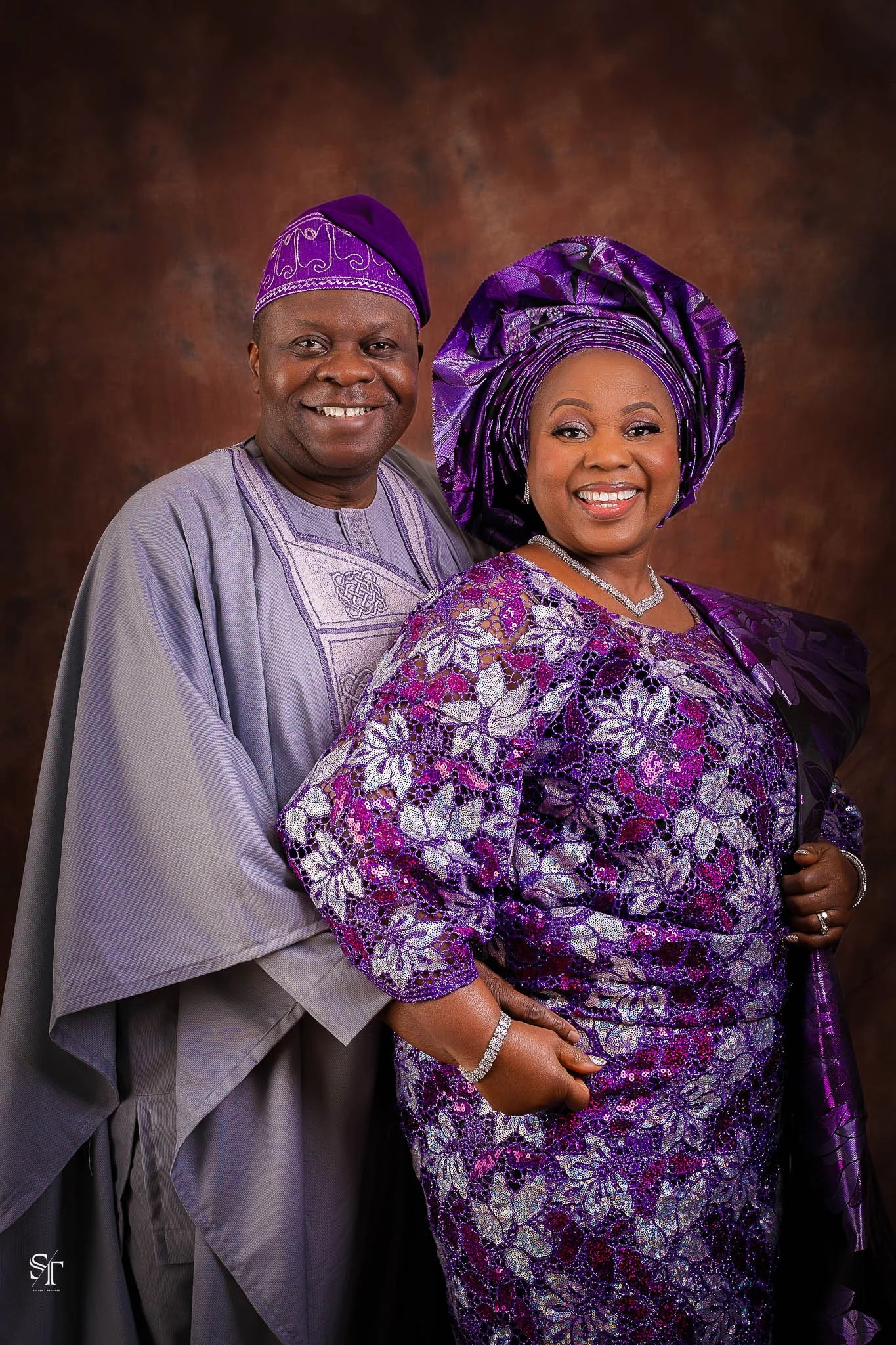 A smiling couple dressed in traditional Nigerian attire, the man in a lavender agbada and purple cap, and the woman in a purple lace dress with matching headwrap, standing against a brown backdrop.