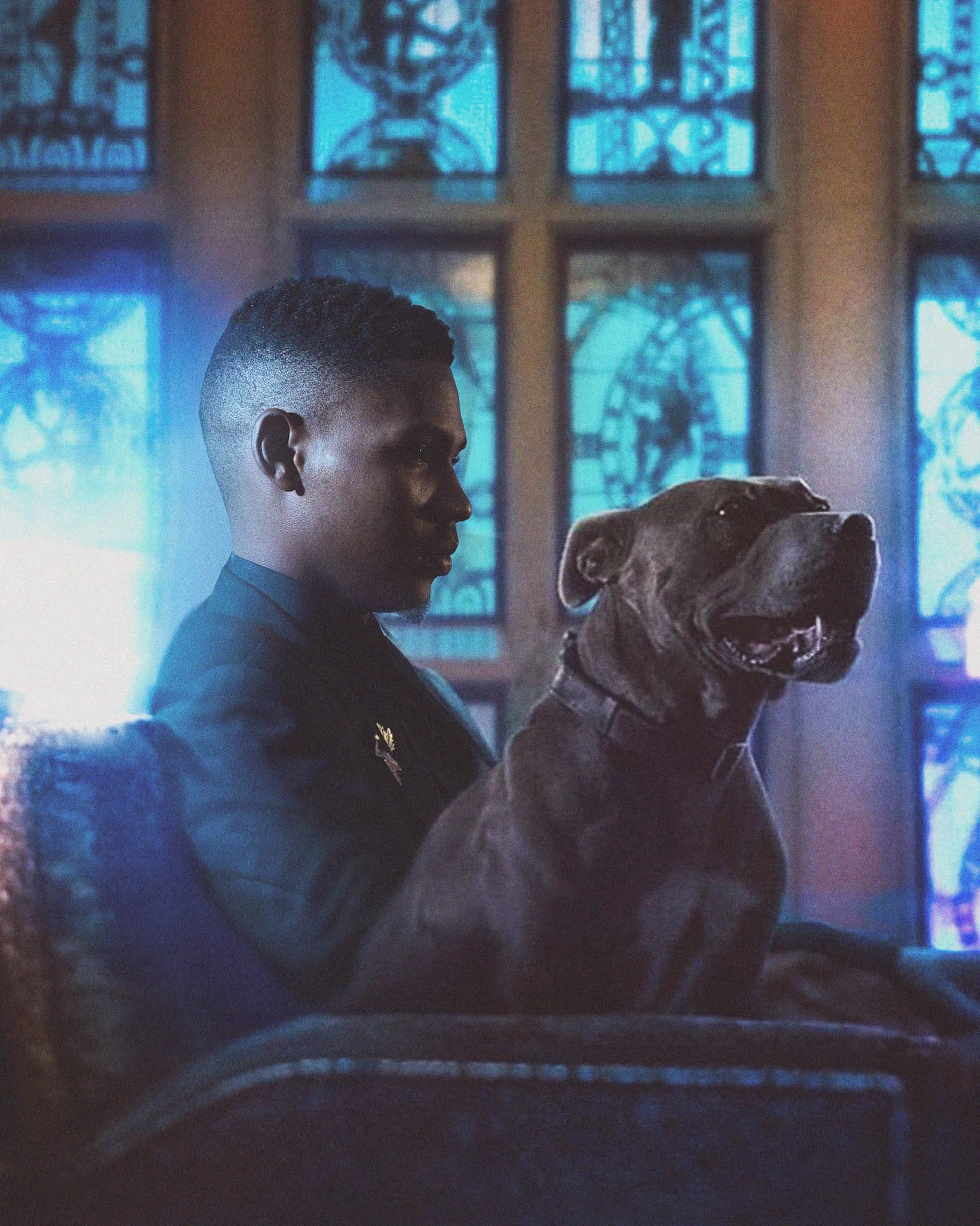 A young man seated indoors with a large dog on his lap, in front of stained glass windows with blue hues.