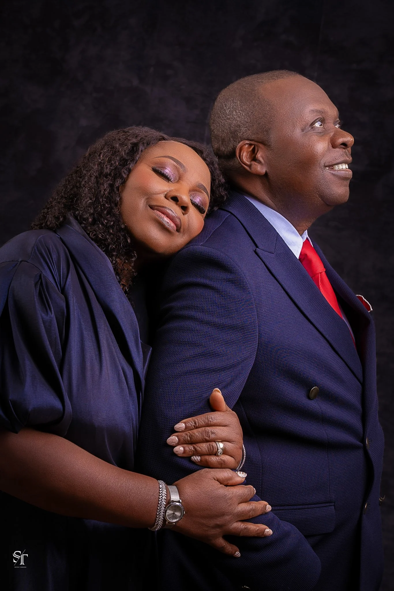 A woman with curly dark hair, wearing makeup and a dark blue dress, leaning her head on the shoulder of a man in a dark blue suit with a red tie, both smiling against a dark background.