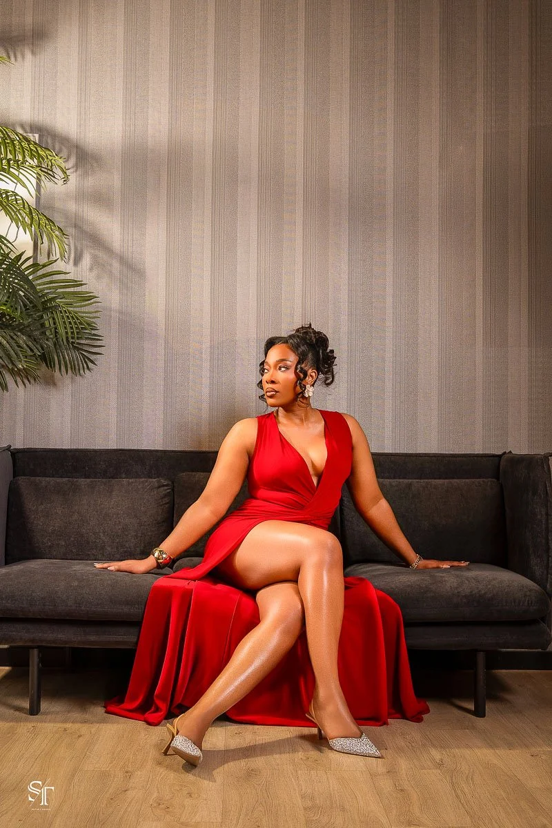 A woman in a red evening gown sits on a black couch in a room with gray striped wallpaper and a potted plant on the left.