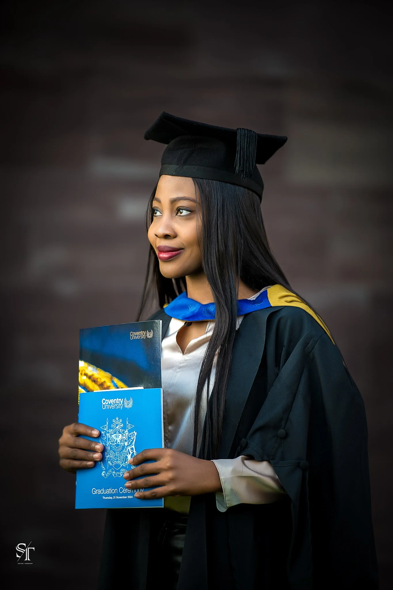 A young woman wearing a graduation cap and gown, holding a blue certificate folder, standing against a dark background.