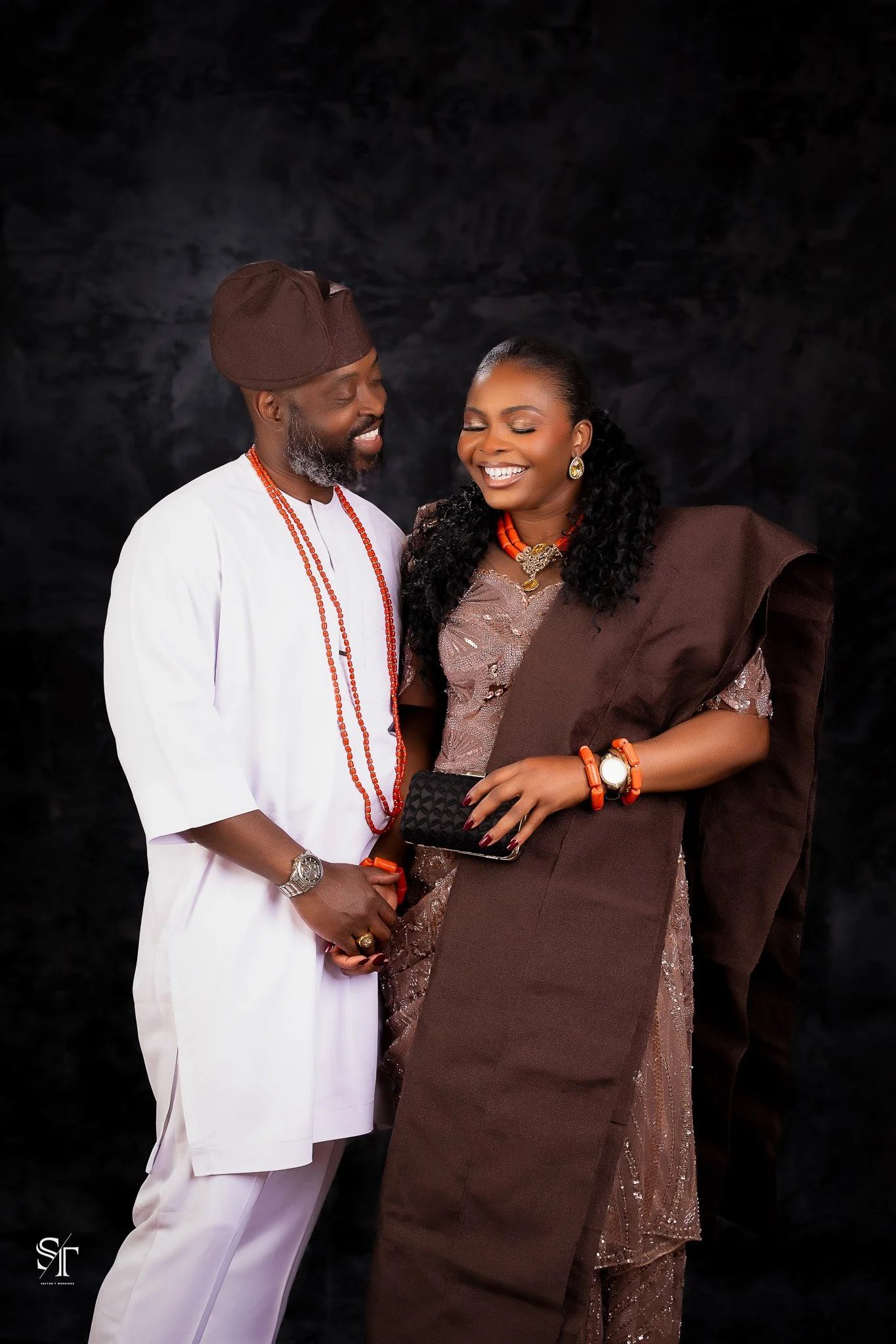 A smiling couple dressed in traditional African attire, holding hands and standing against a black background.