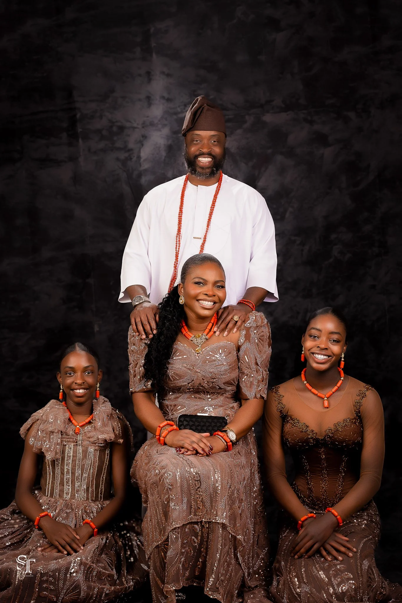 A family of four dressed in traditional African attire posing for a studio portrait against a dark background. The man is standing behind the woman and children, wearing a white garment, brown cap, and coral bead necklace. The woman and two young gir