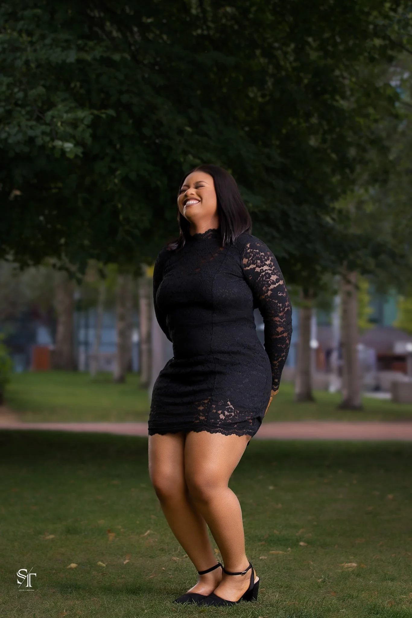 A woman in a black lace dress and black heels standing outdoors on grass, smiling with eyes closed, in front of trees.