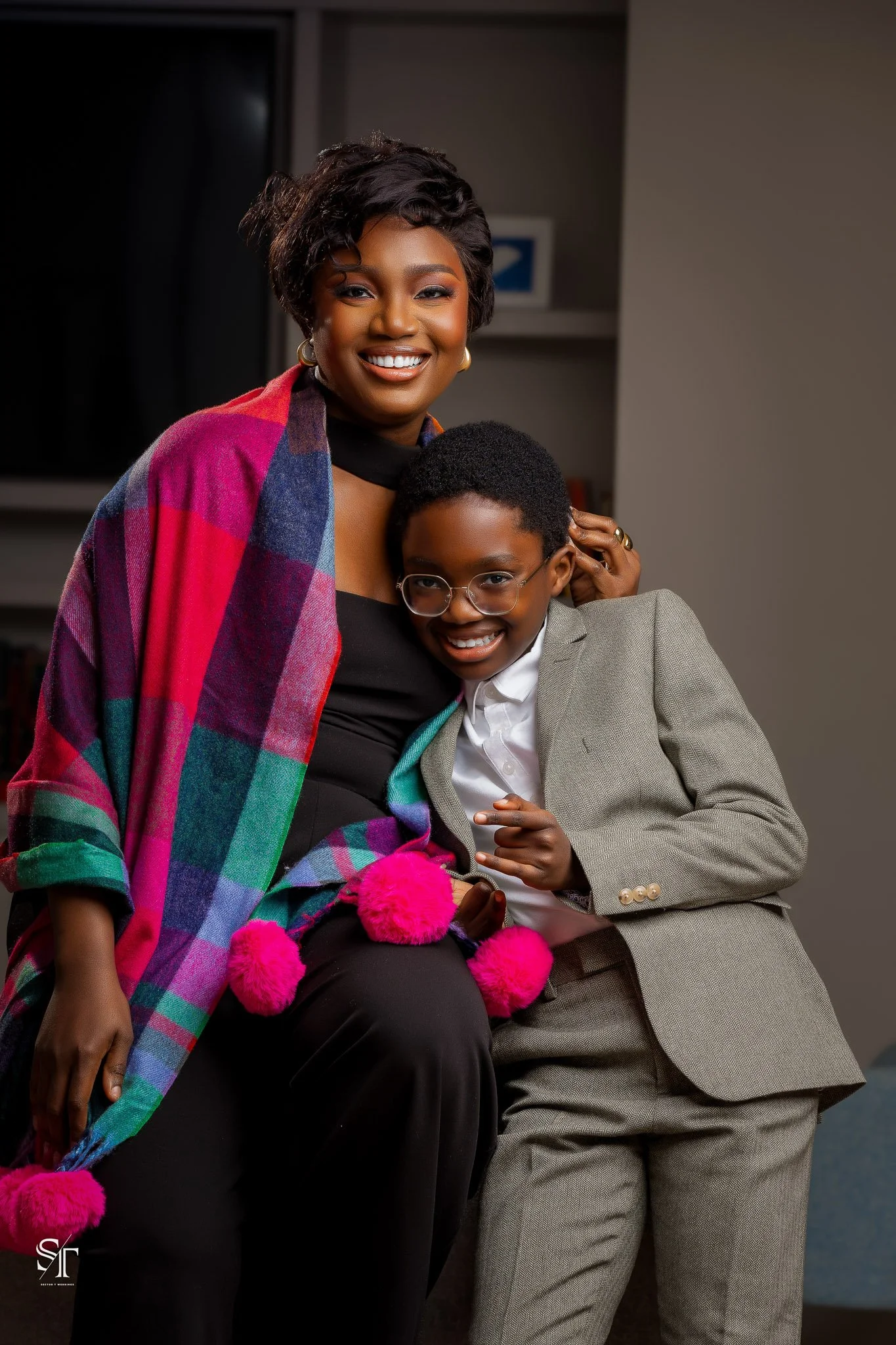 An adult woman and a young boy, both smiling, standing close together in an indoor setting. The woman is wearing a colorful plaid shawl, black top, and gold earrings. The boy is dressed in a gray suit with a white shirt and glasses.