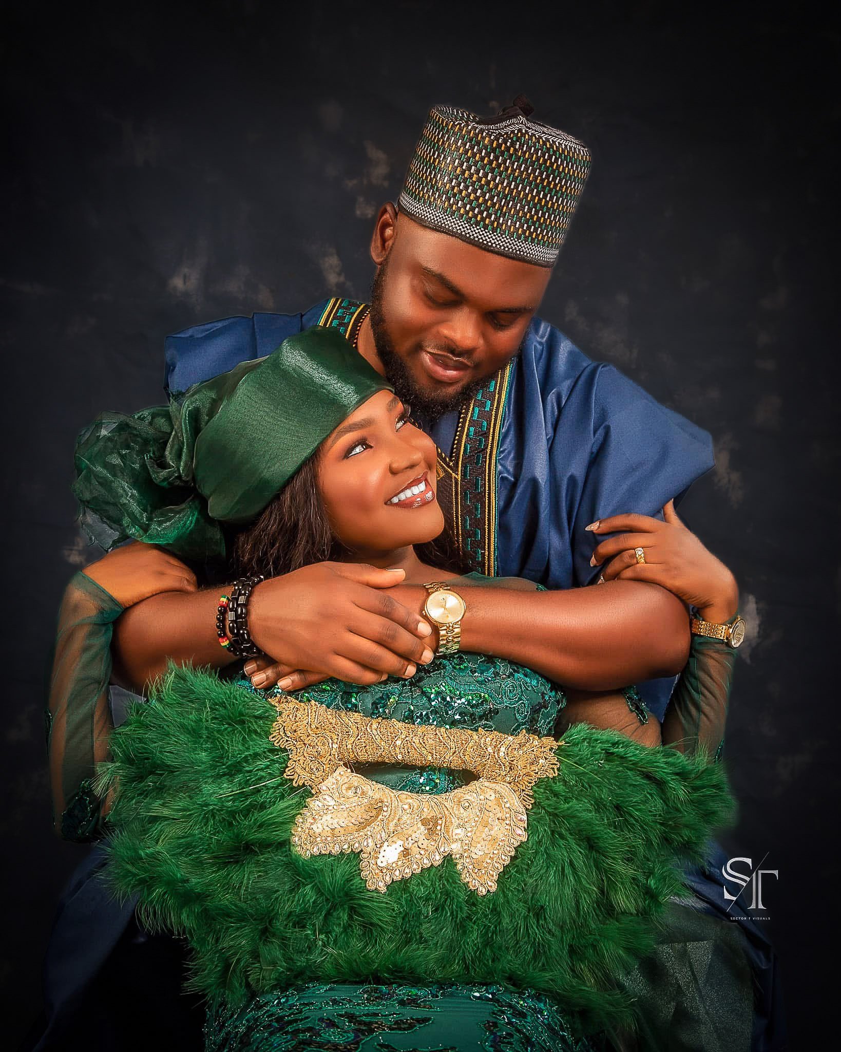 A man and woman dressed in traditional Nigerian attire embrace, smiling, against a dark background.