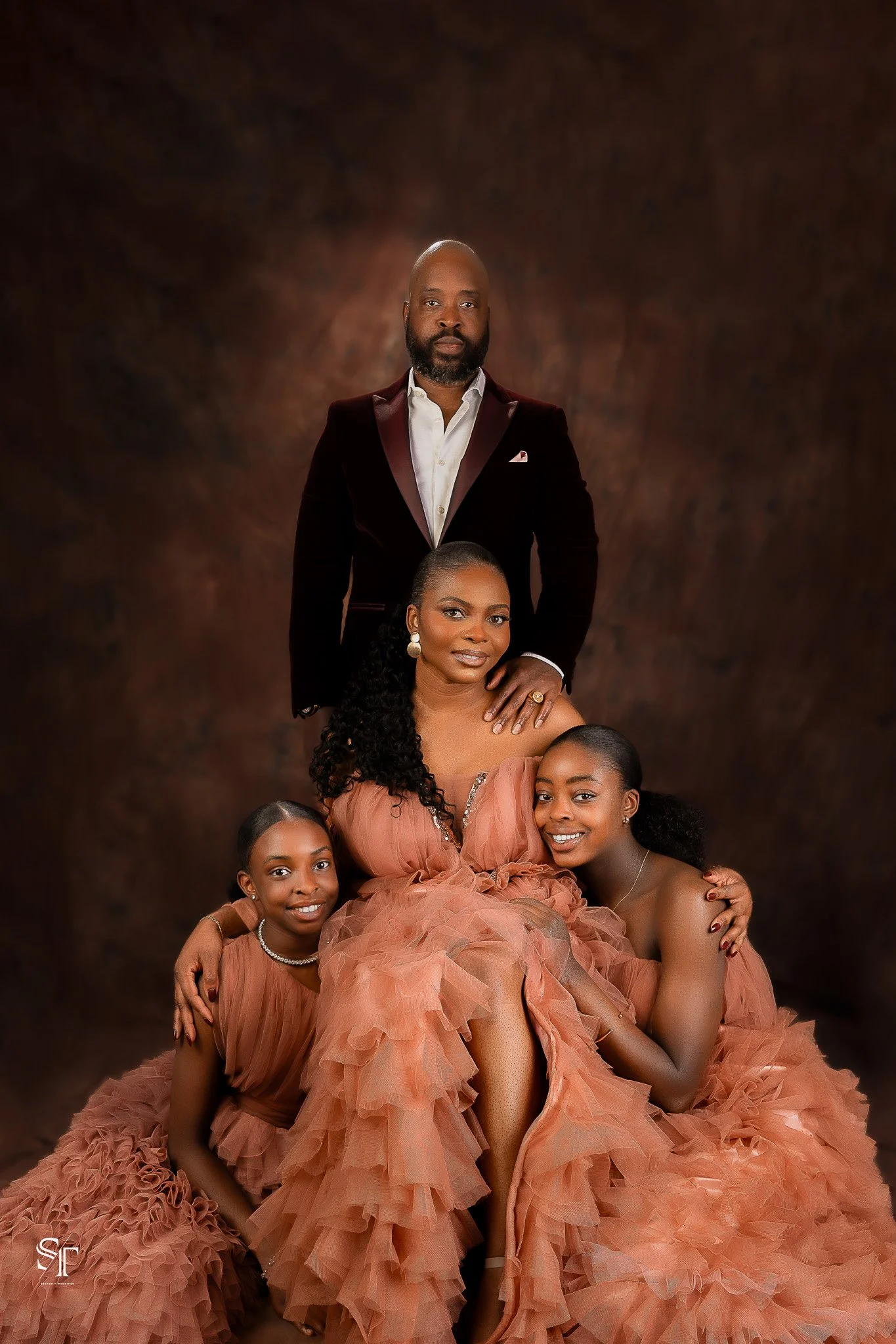 A family portrait featuring a man in a dark tuxedo with a white shirt and three women in matching peach ruffled dresses, posed against a dark, textured background.