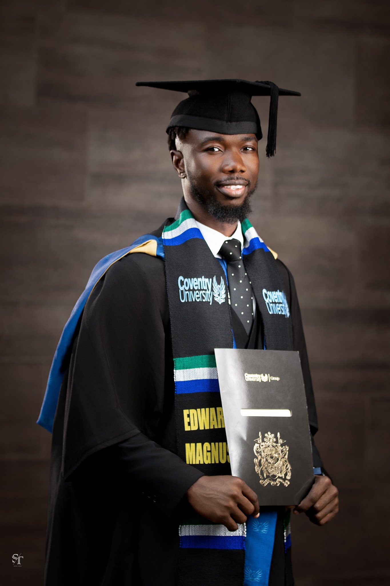 Graduation portrait of a young man of African descent wearing a cap and gown with a colorful stole, holding a diploma, standing against a neutral background.