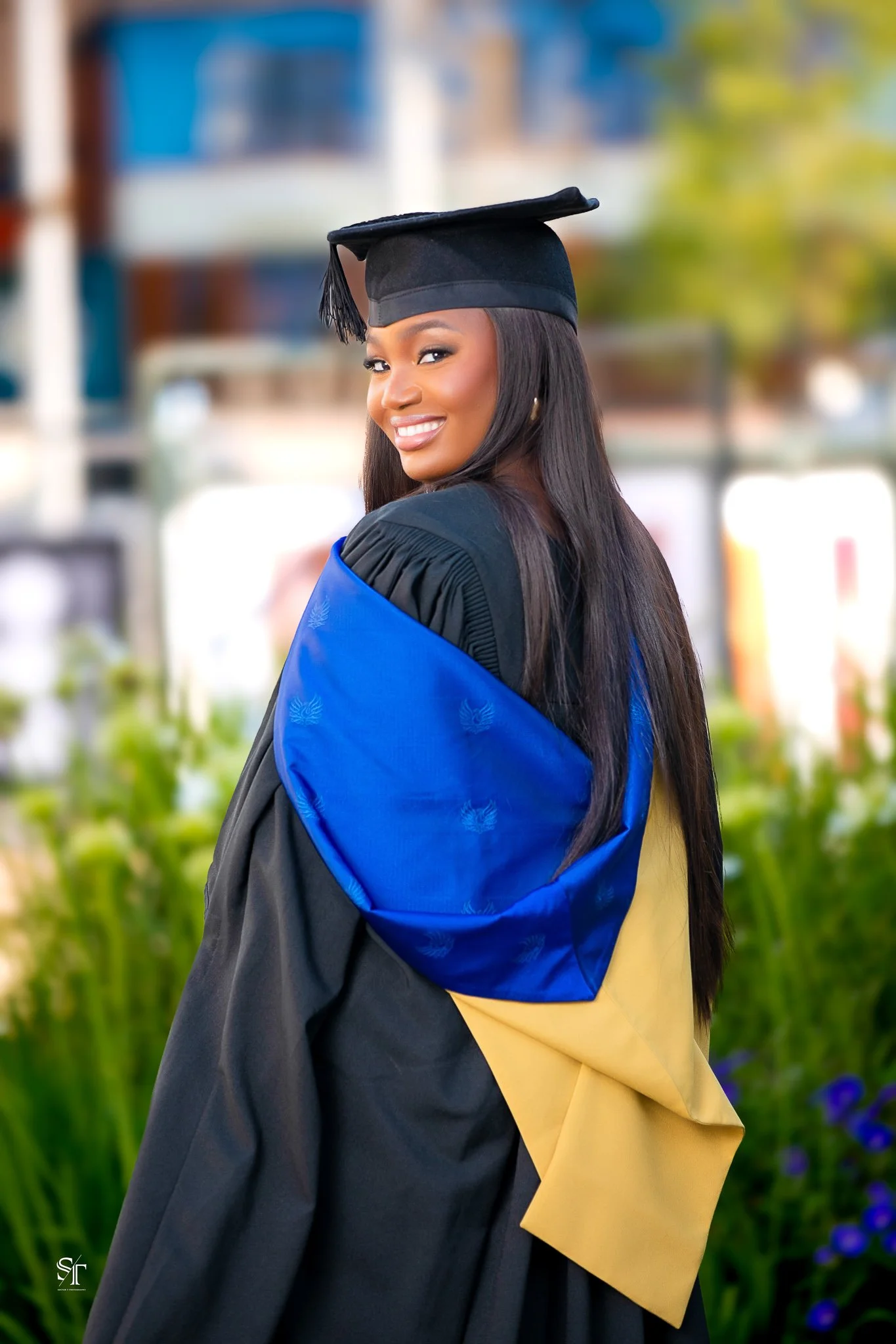 A young woman in graduation attire, smiling and looking over her shoulder, standing outdoors with a blurred background of greenery and buildings.