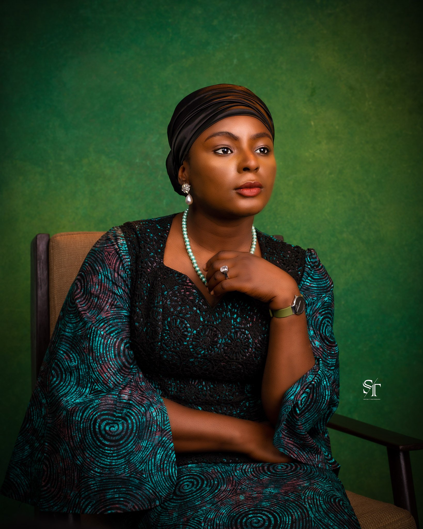 Portrait of a woman with dark hair wrapped in a black headscarf, wearing pearl earrings and necklace, a ring, a wristwatch, and a patterned dress with teal and black swirls, seated with her hand near her face against a green background.
