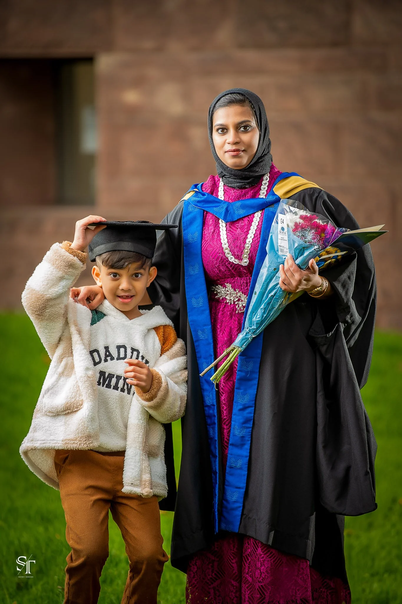 A woman in a cap and gown celebrating her graduation with a young boy in a white hoodie. The woman holds a bouquet of flowers and a diploma, standing outside on grass with a brick building in the background.