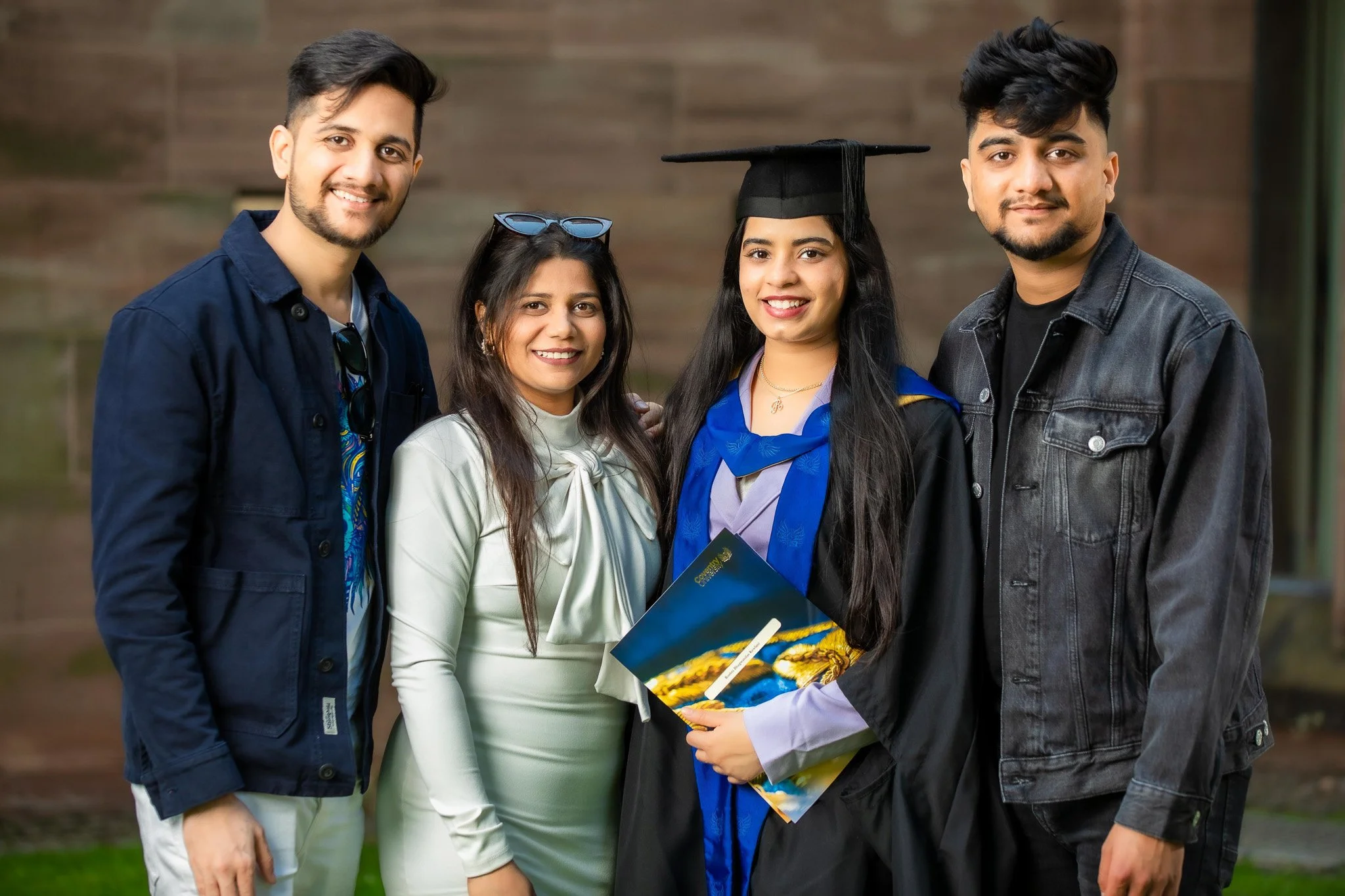 Group of five people celebrating graduation outdoors, with a woman wearing a graduation cap and gown holding a diploma, smiling.