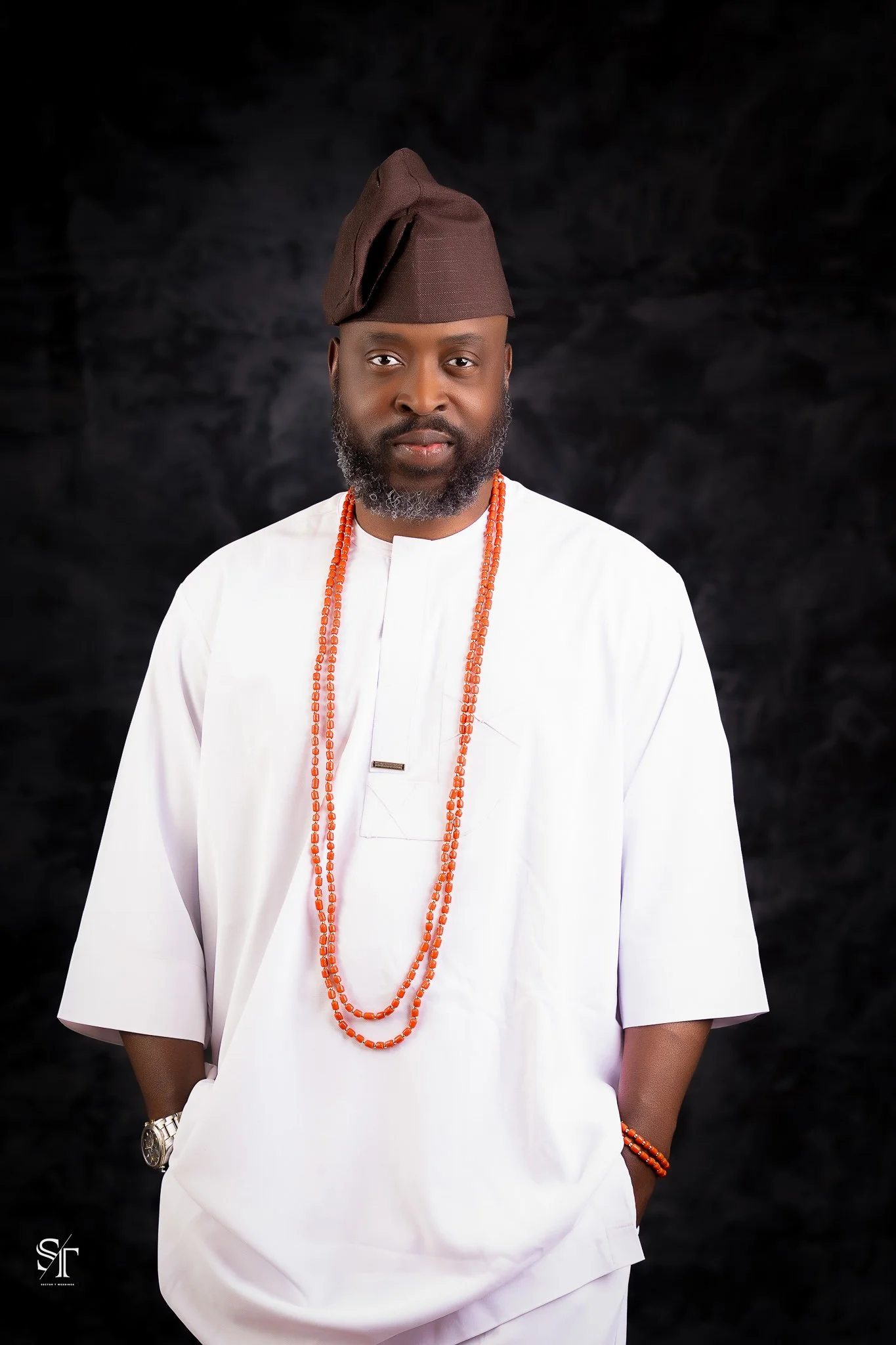 A man dressed in traditional Nigerian attire, including a white agbada, multiple beaded necklaces, matching beads on his wrist, a watch, and a brown cap, standing against a dark background.