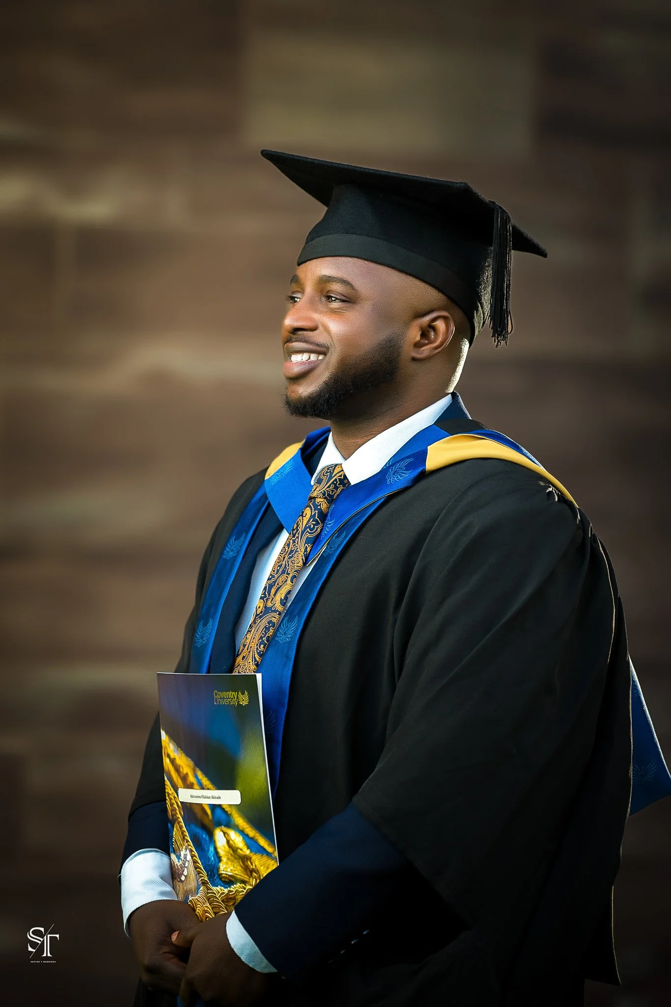 A smiling man in graduation cap and gown holding a diploma in front of a brown blurred background.