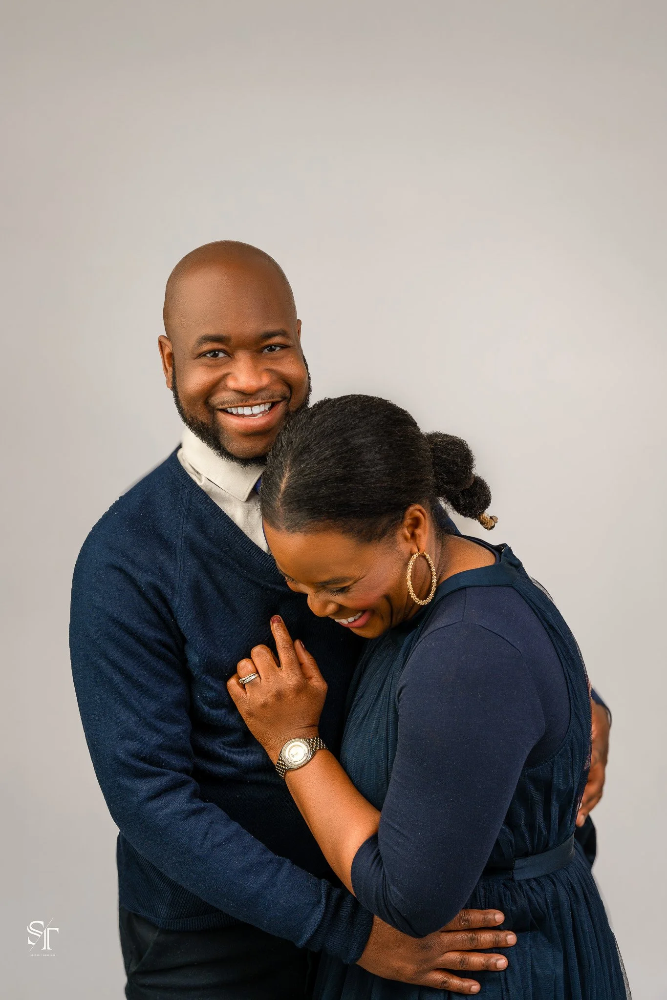 A joyful man and woman sharing a hug, both smiling, with the woman leaning her head on the man's chest. They are dressed in dark blue tops, and the background is plain and light-colored.