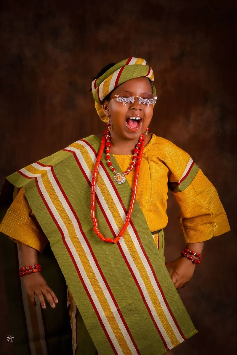 A young girl dressed in traditional African attire with vibrant yellow and green fabric, accessorized with red beads, jewelry, and a head wrap, smiling joyfully with her eyes closed.