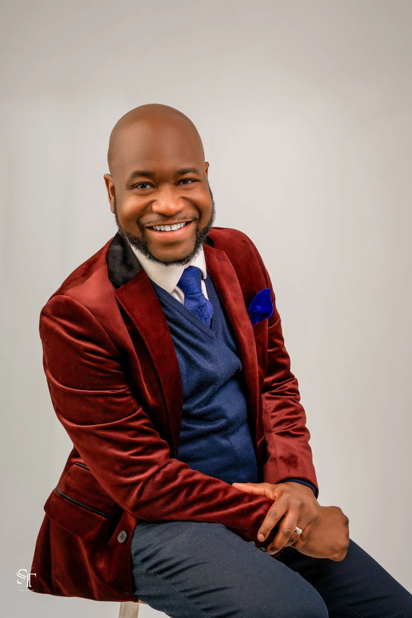 A smiling man in a red velvet blazer, navy sweater, and white shirt with a blue tie, sitting against a plain light-colored background.