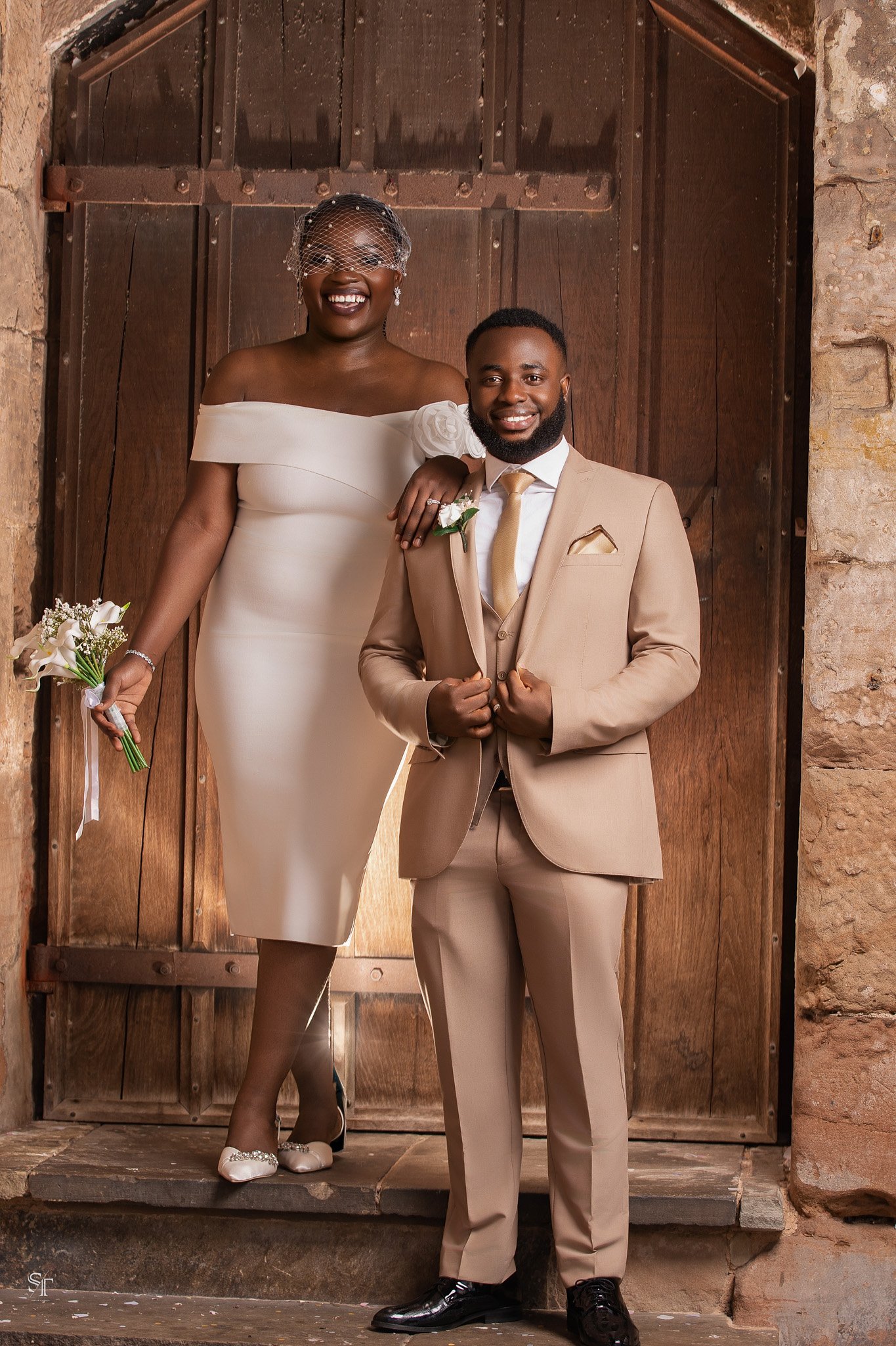 A bride and groom standing inside a rustic stone and wood doorway. The bride is wearing a white off-shoulder wedding dress with a veil, and holding a small bouquet. The groom is wearing a beige suit with a white shirt, gold tie, and black shoes.