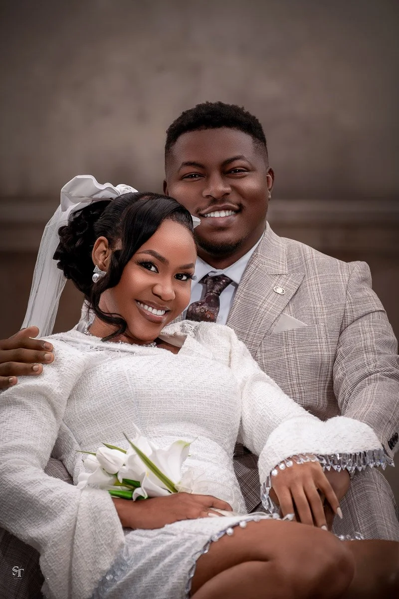 A smiling couple dressed in wedding attire, with the woman holding white calla lilies, posing indoors against a plain background.