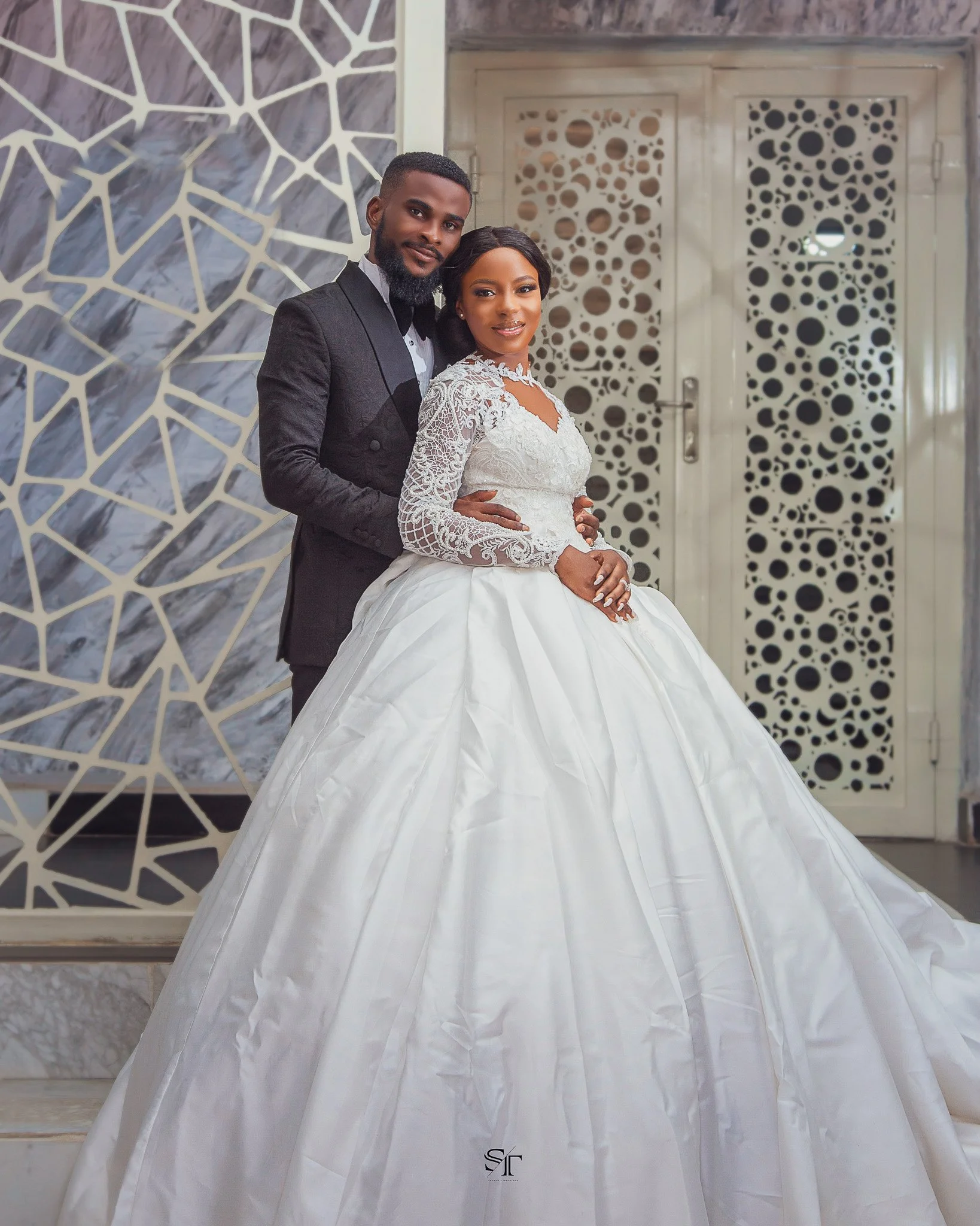 A bride and groom in wedding attire posing indoors. The bride wears a white wedding dress with lace details and a full skirt, while the groom wears a black suit with a gray shirt and black bow tie. They stand close together with the groom holding the