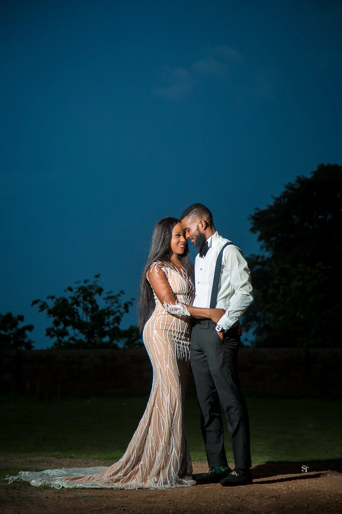 A couple dressed elegantly, standing close together outdoors at dusk, with their foreheads touching and smiling.
