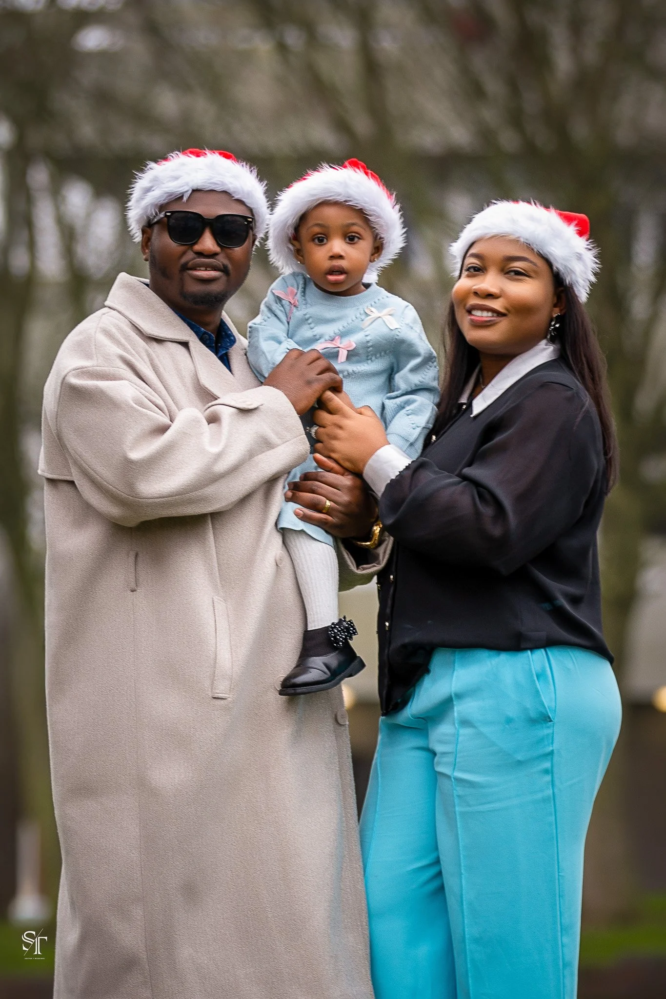 Family of three wearing Santa hats, outdoors with blurred trees in background. Man with sunglasses holding young girl, woman standing beside them.