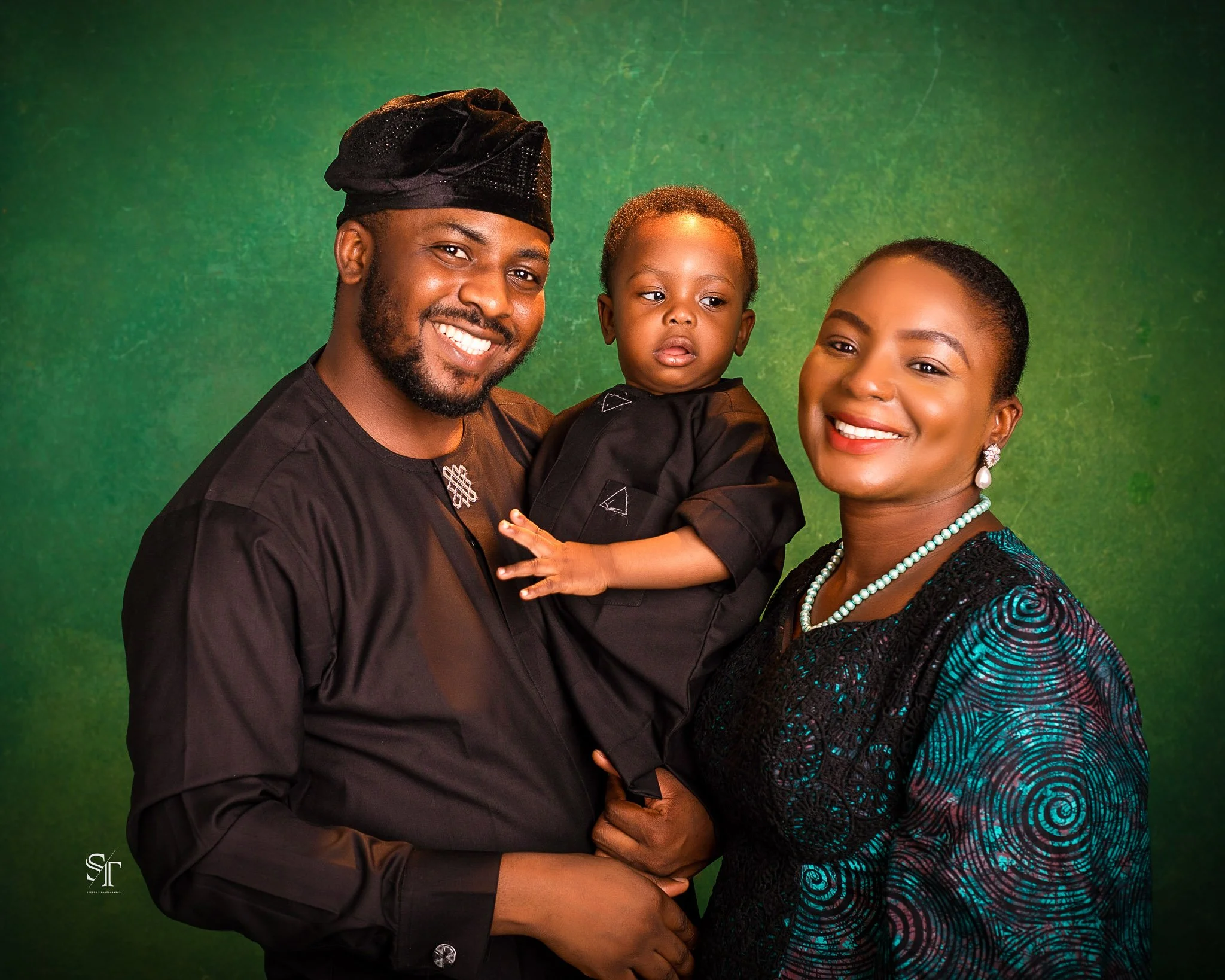 A smiling black family of three with a father, mother, and young child dressed in black, standing against a green background.