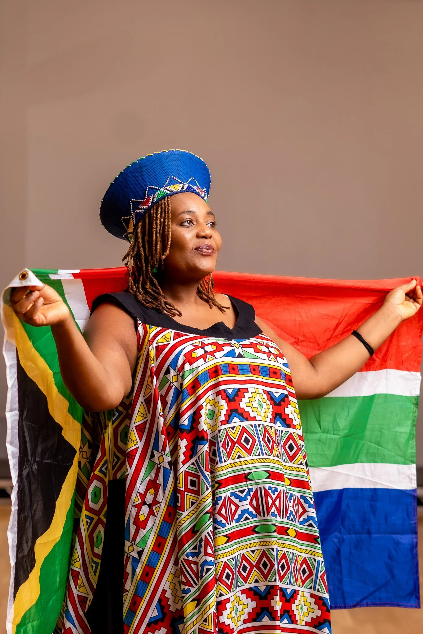 A woman wearing traditional African attire and a colorful beaded hat, holding a South African flag.
