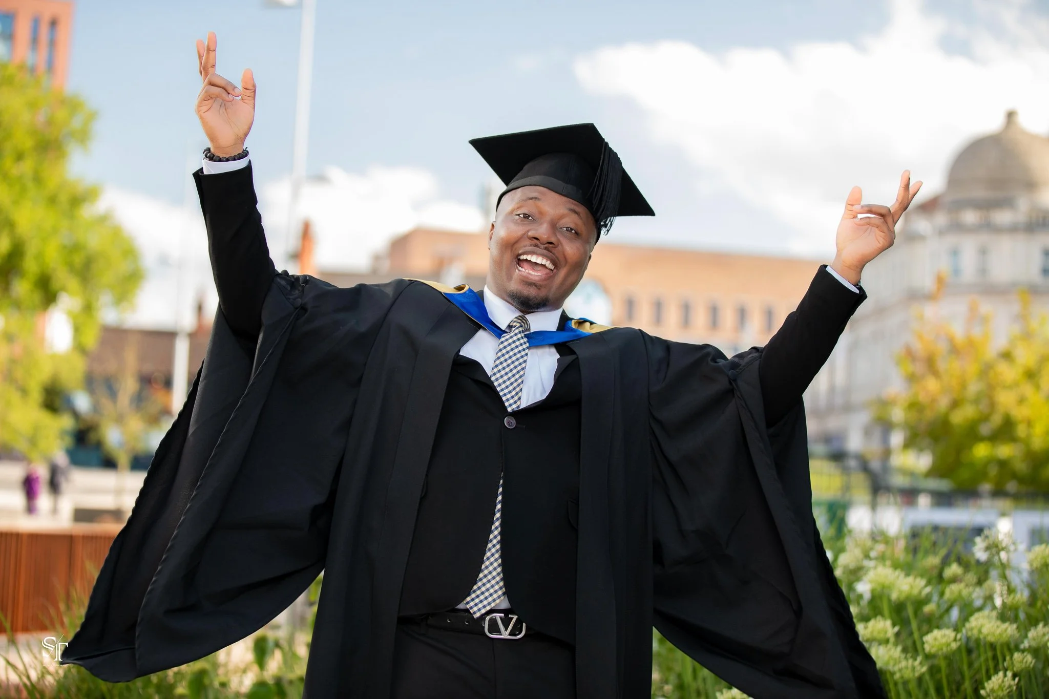 A man in a graduation cap and gown celebrating outdoors with arms raised, smiling with a background of trees and buildings.