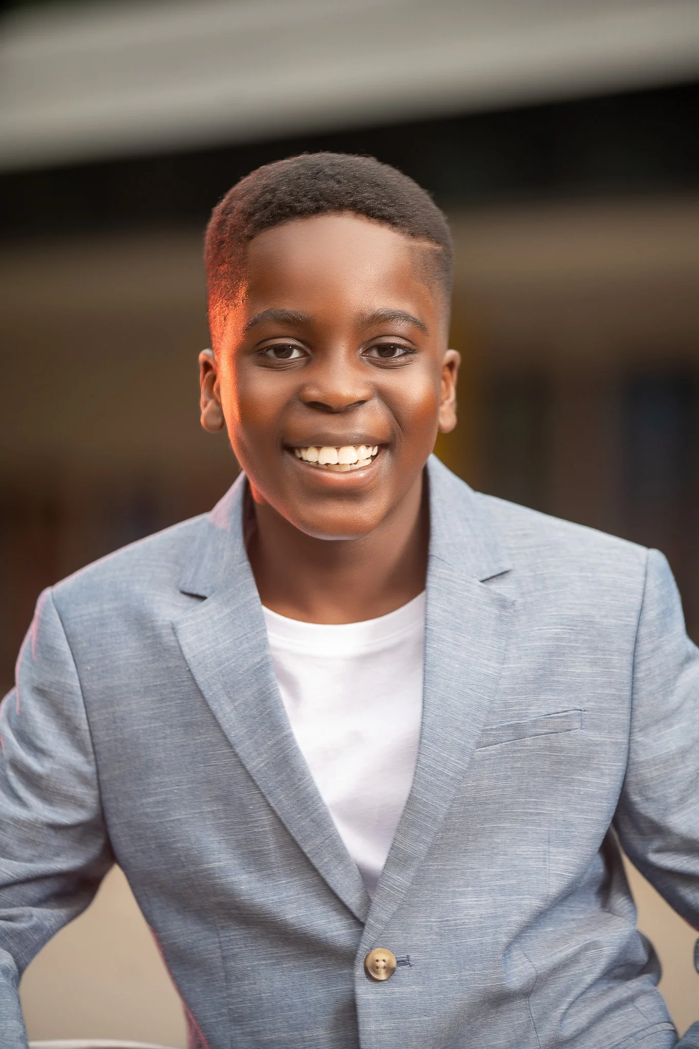 Smiling young boy in a light gray blazer and white shirt, outside during sunset.