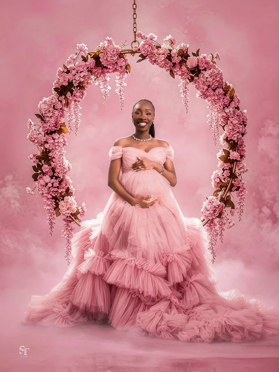 A pregnant woman wearing a pink, ruffled, off-the-shoulder gown stands in front of a pink floral arch, smiling and holding her belly in a studio setting.