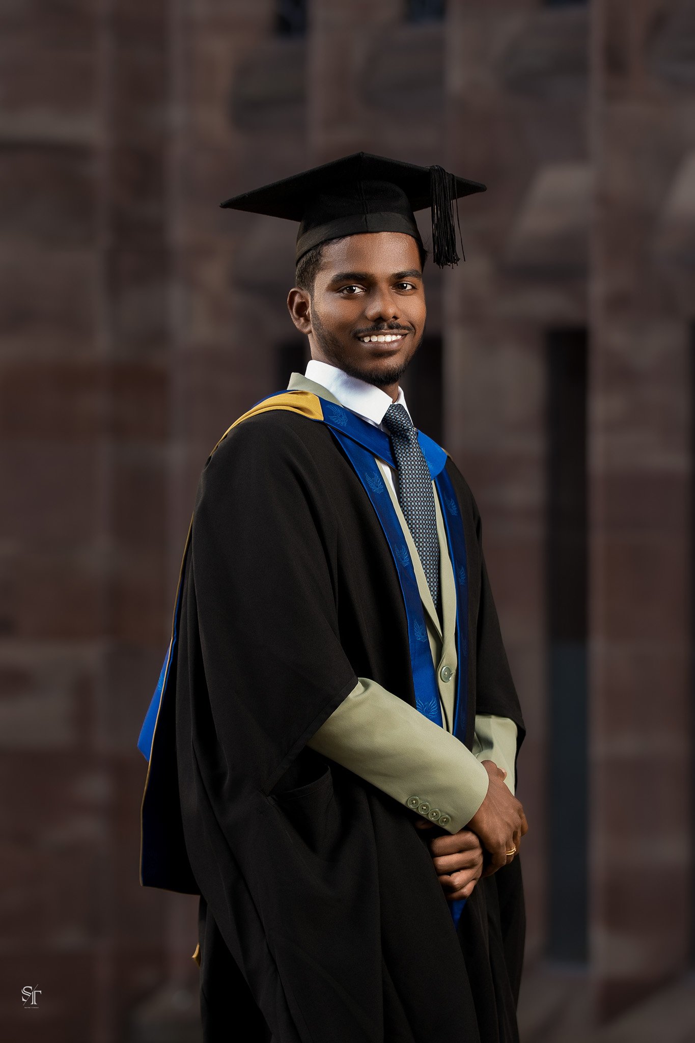Young man in graduation cap and gown smiling, standing in front of a blurred dark background.