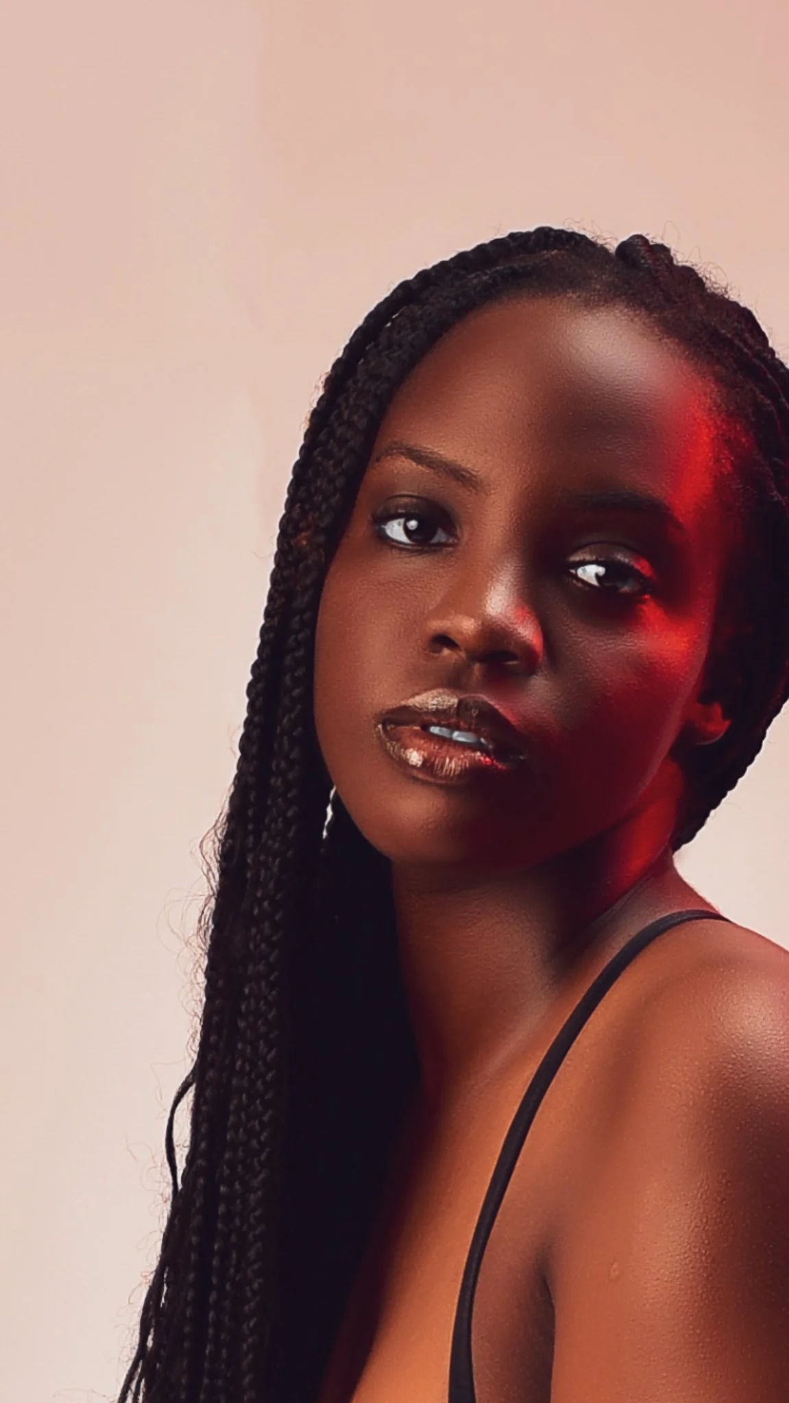 A young woman with dark skin and long braided hair, wearing a black spaghetti strap top, looking at the camera with a neutral expression against a plain light-colored background.