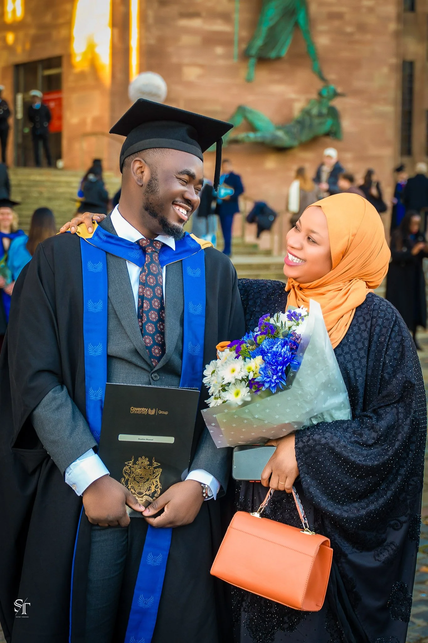 A graduation ceremony where a male graduate wearing academic cap and gown is smiling and embracing a woman with a bouquet of flowers, with other graduates and attendees in the background.