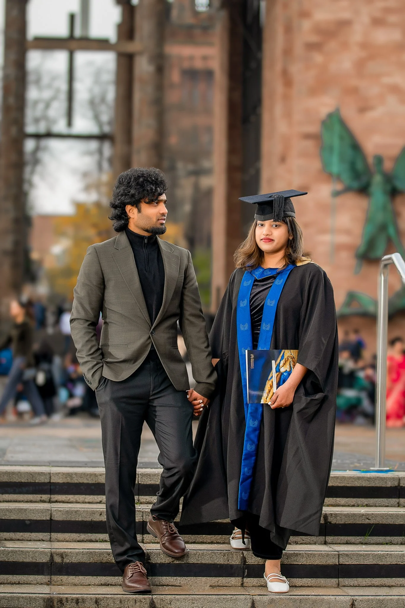 A woman in graduation gown and cap holding a diploma, standing on steps next to a man in a suit, holding her hand, outdoors during daytime.