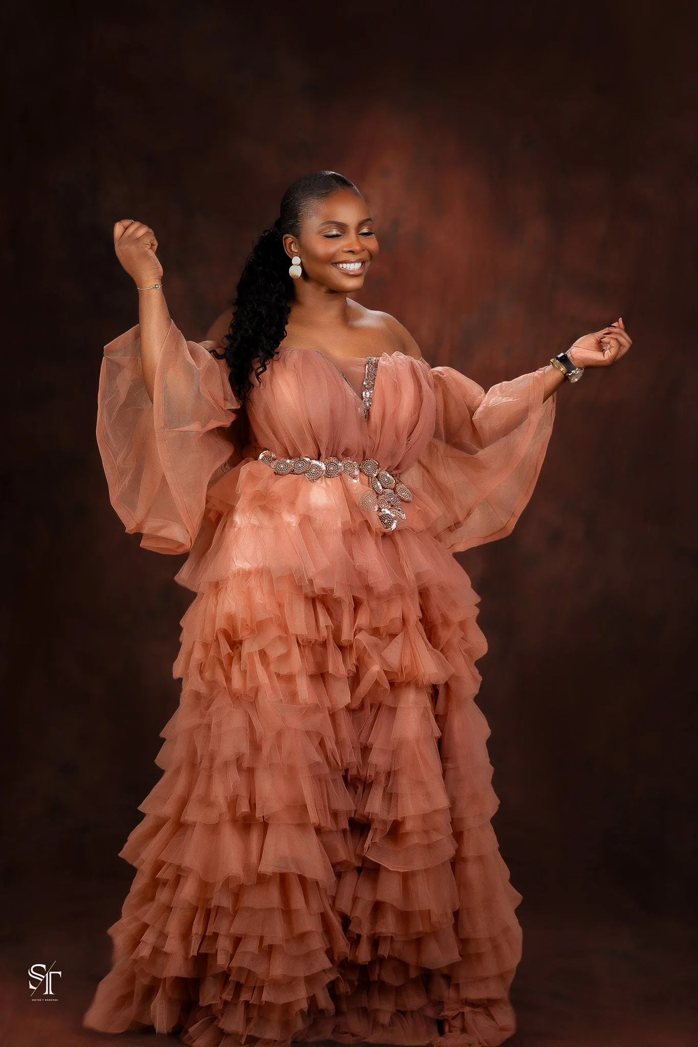 Woman smiling and posing in a ruffled, peach-colored gown with sheer sleeves and decorative belt against a dark brown background.