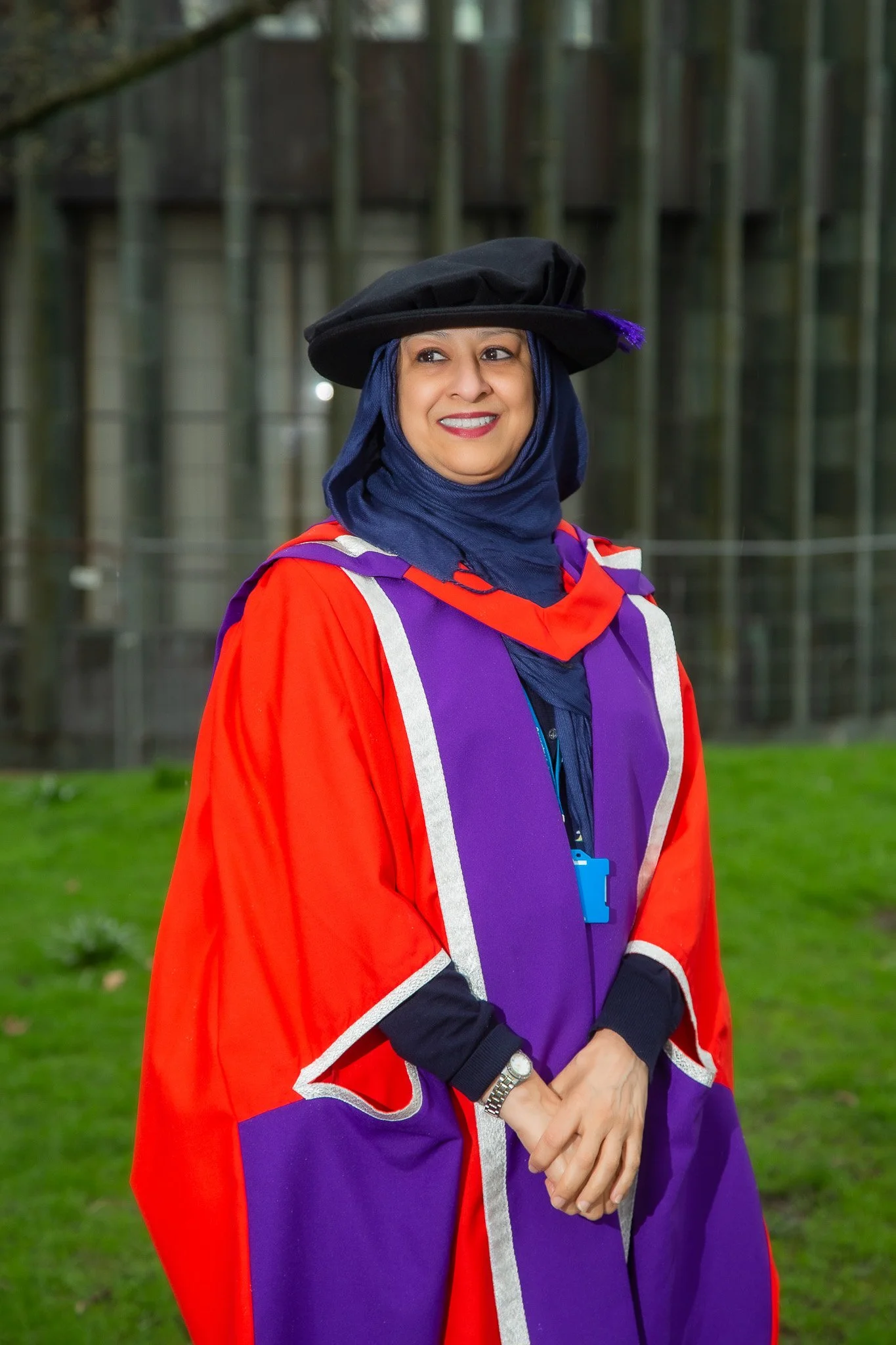 Woman in a graduation gown and cap outdoors with a background of trees and a building.