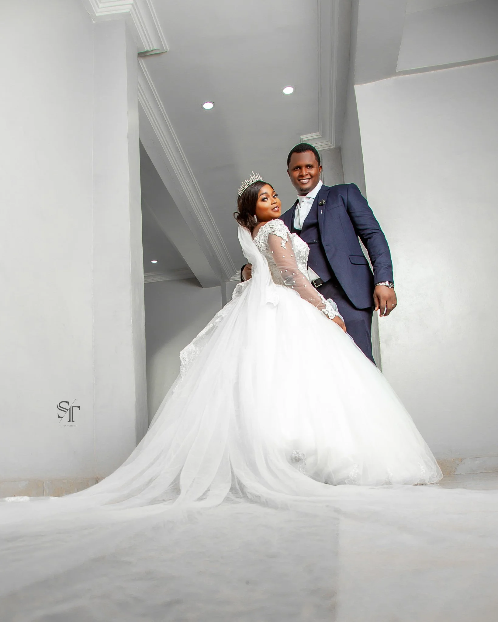 A newlywed couple dressed in wedding attire, with the bride in a white gown and tiara, and the groom in a dark blue suit, standing indoors against gray walls.