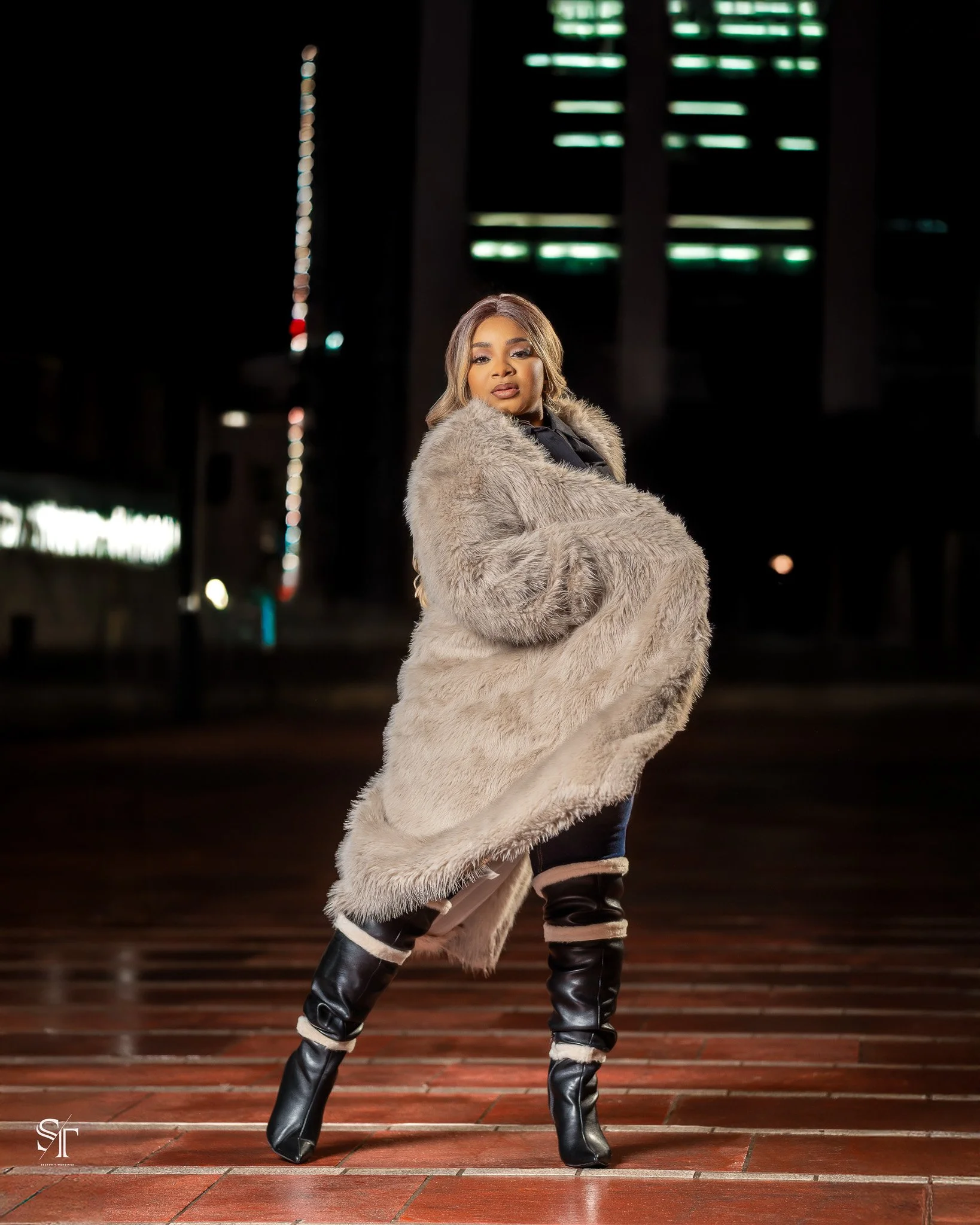 A woman standing on a brick street at night, wearing a long beige fur coat, black boots with beige accents, and posing confidently with a cityscape background and illuminated buildings.