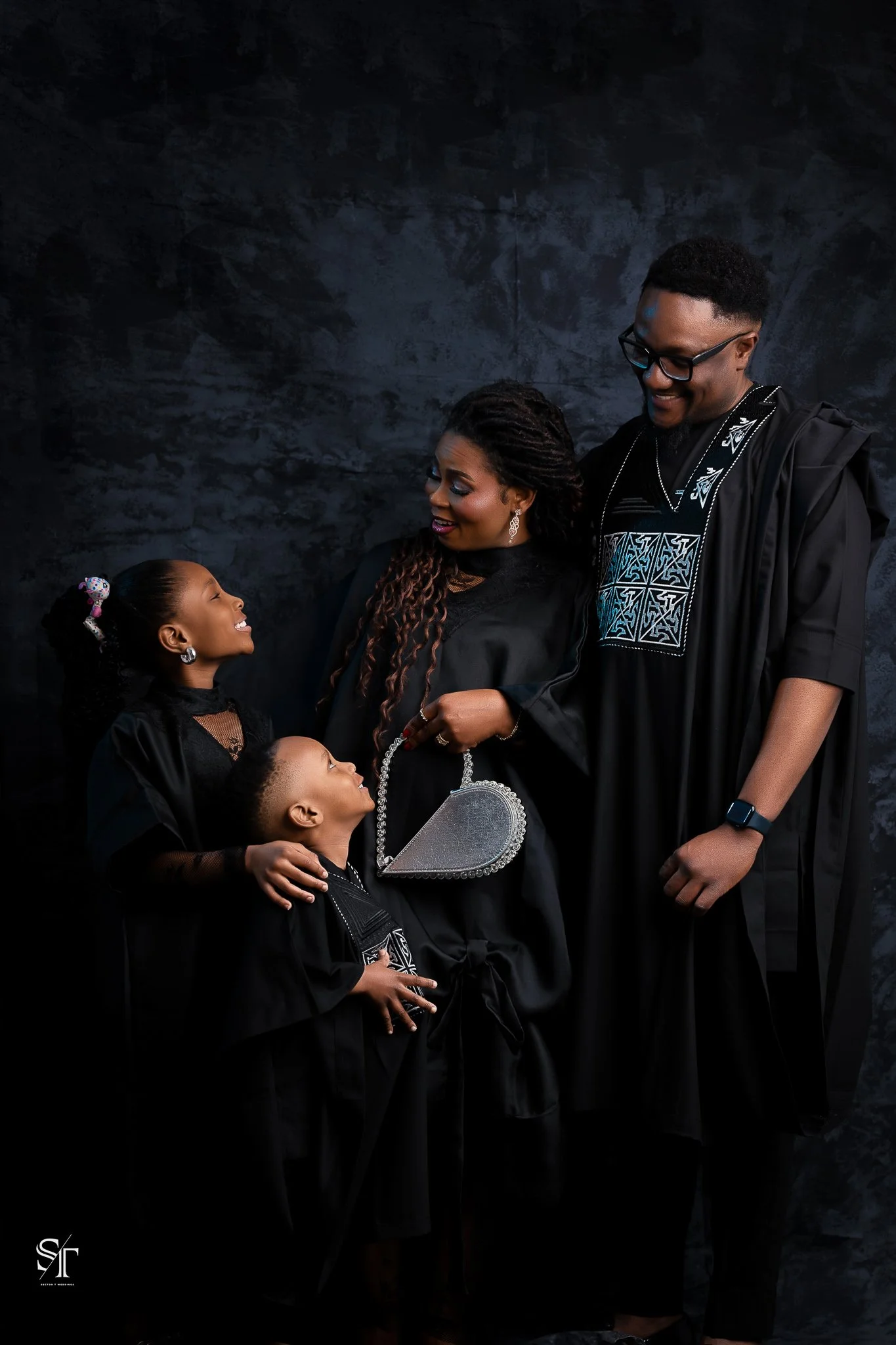 A family celebrating a graduation, dressed in black gowns. The mother holds a silver heart-shaped purse, and the father wears a smartwatch. Two children stand beside them, smiling and looking at their parents.