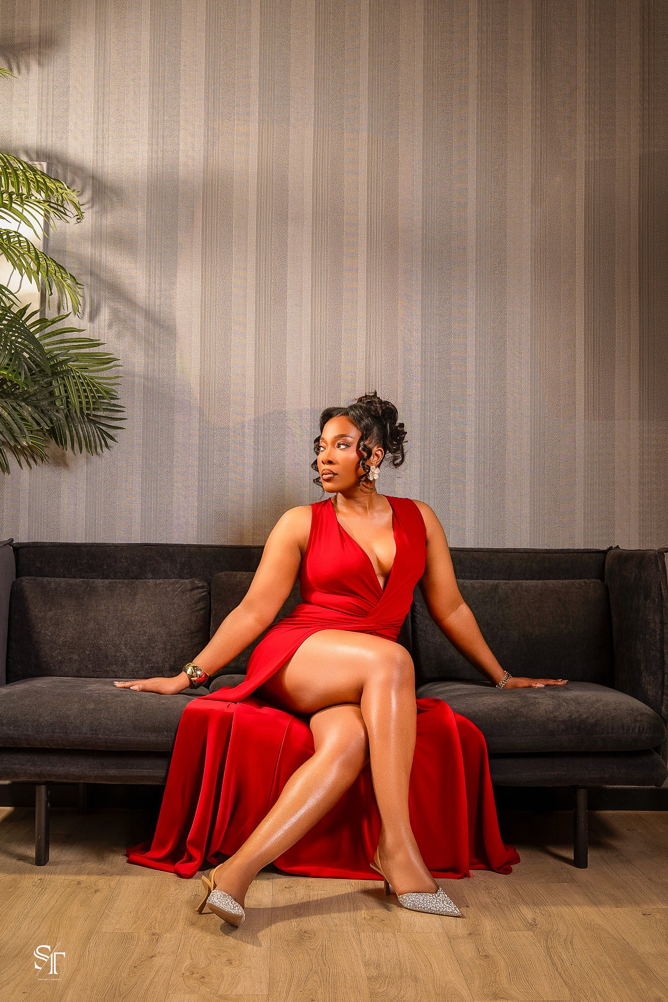 A woman in a red dress sitting on a dark sofa with one leg crossed over the other, wearing high heels, earrings, bracelet, and watch, posed in front of a textured gray wall and a potted plant.