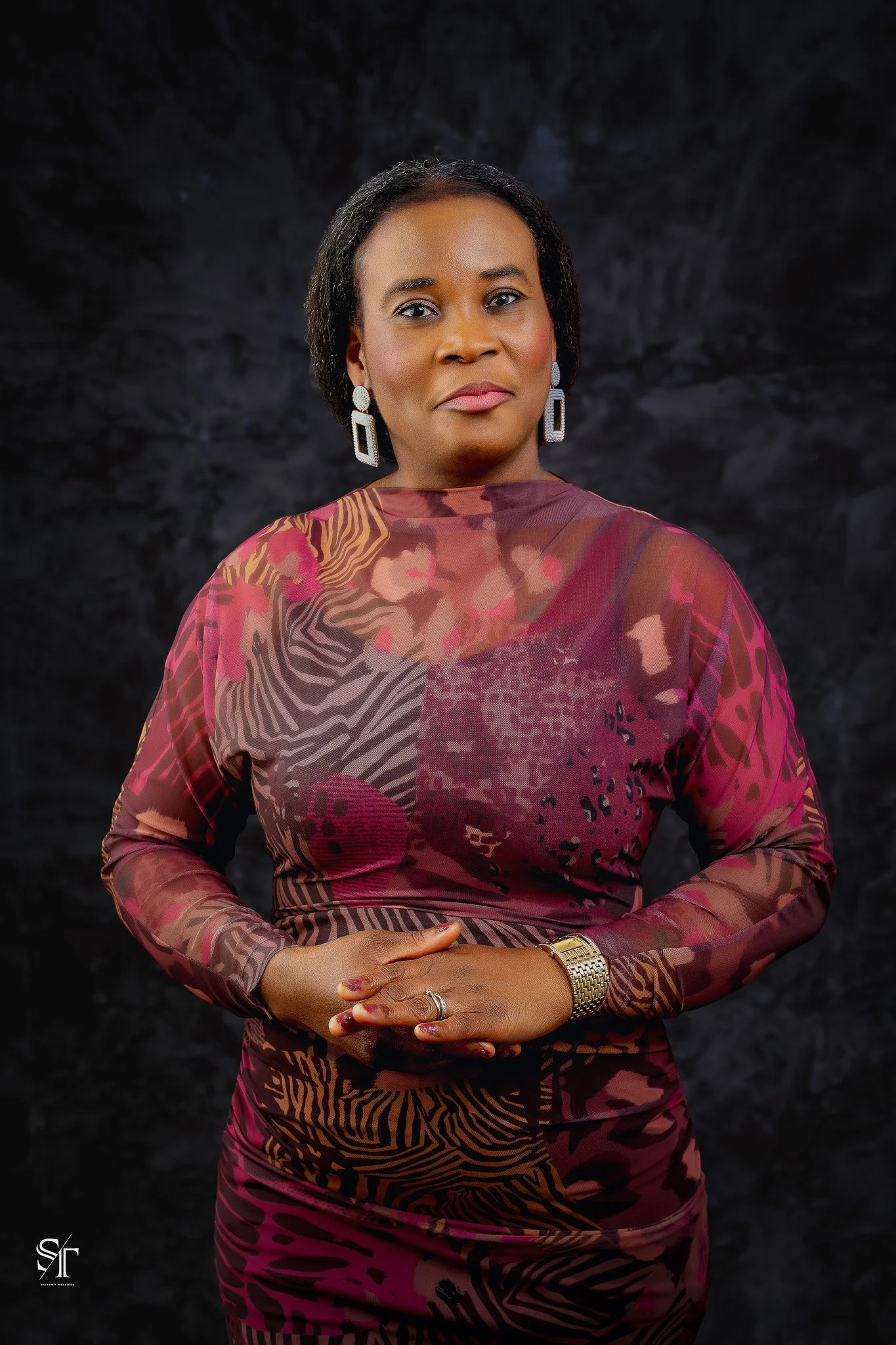 A woman with short black hair, wearing large earrings, a colorful patterned dress in shades of maroon, pink, and brown, with a gold watch on her left wrist, standing against a dark background.