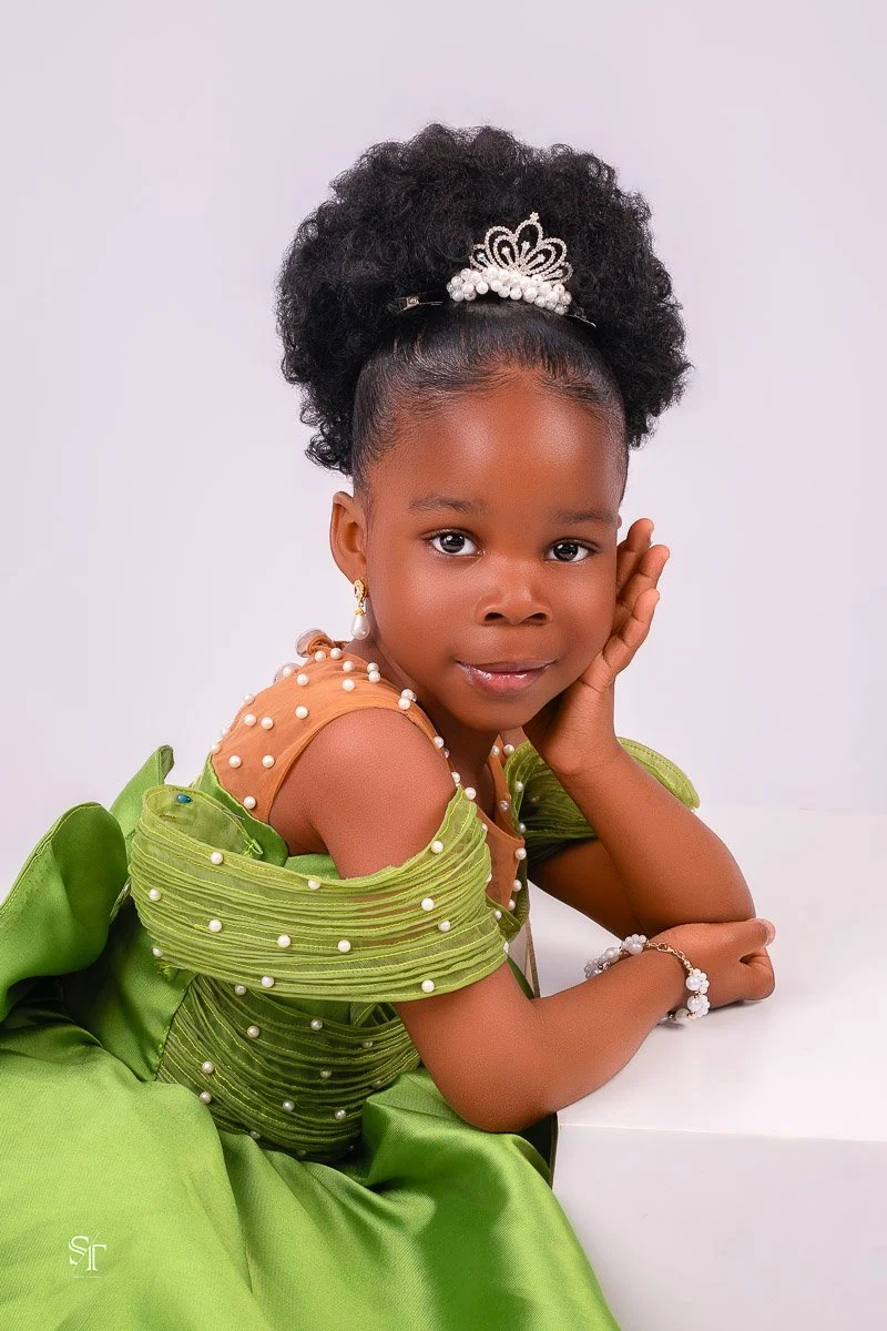 A young girl in a green dress with pearl embellishments, wearing pearl jewelry and a tiara, poses with her hand resting on her cheek against a plain white background.
